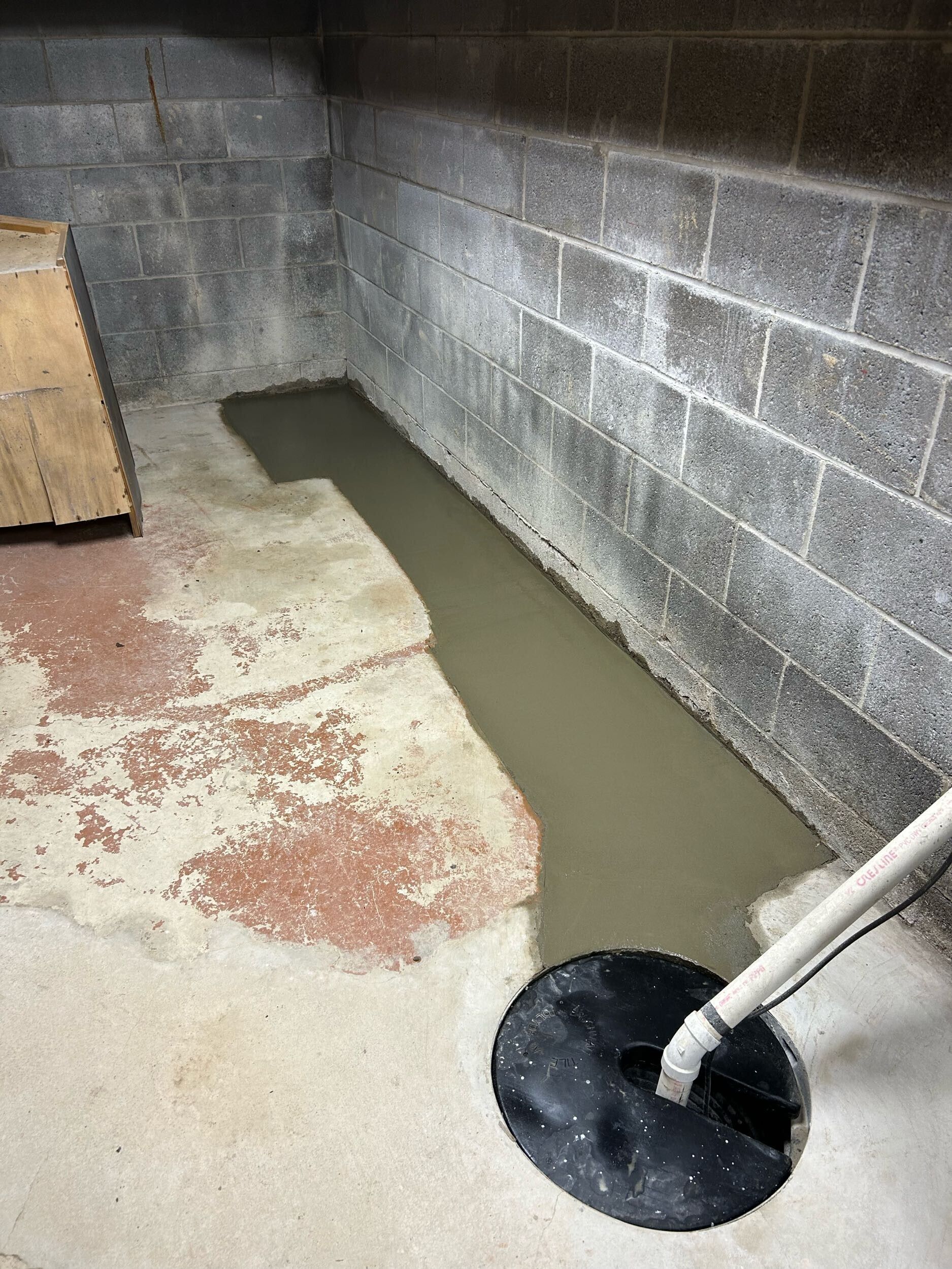 Concrete crawlspace with a narrow trench of wet cement beside a cinder block wall and drain pipe