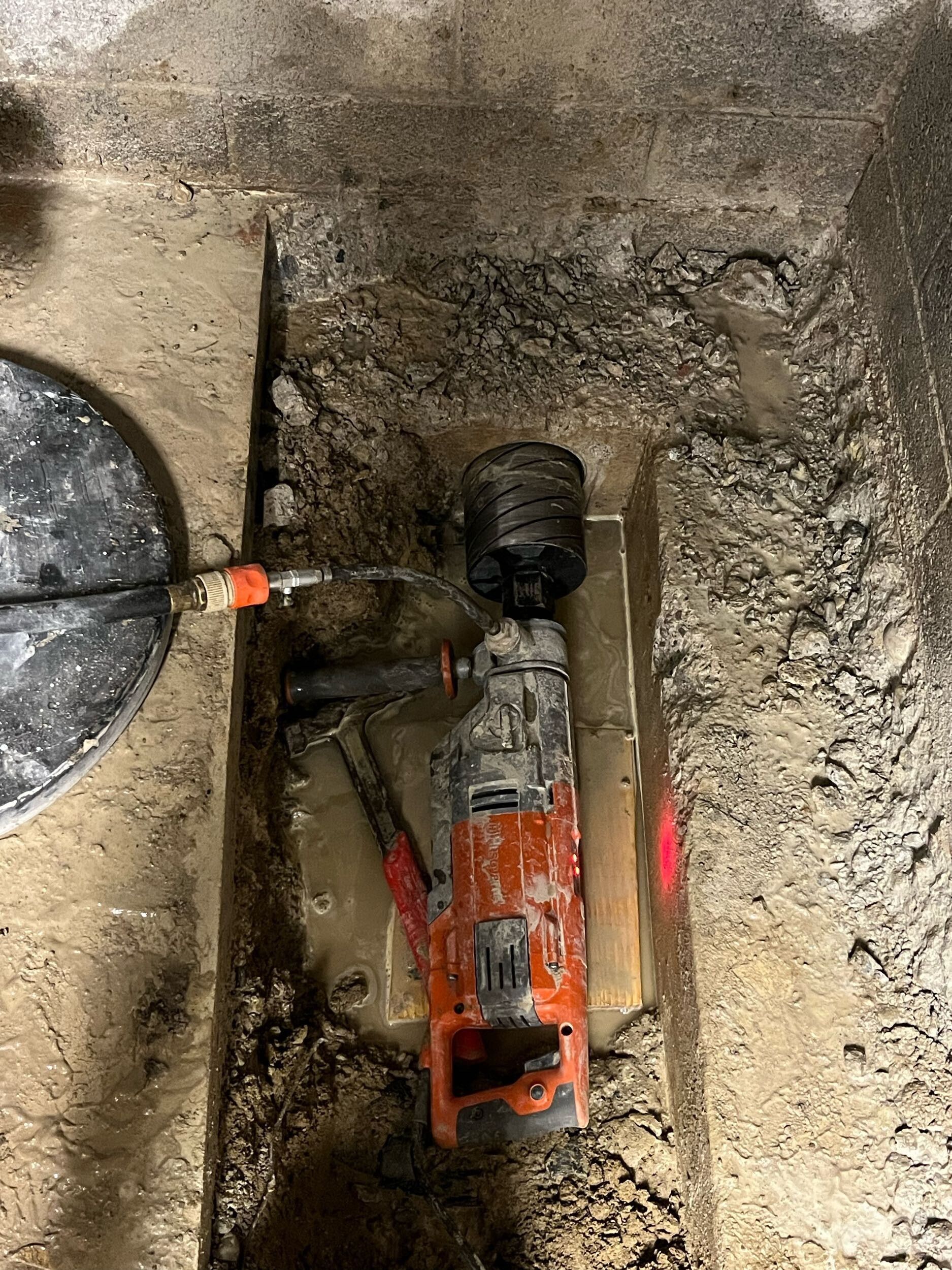Orange power tool in a dirt-lined trench beside a shovel and black pipe.