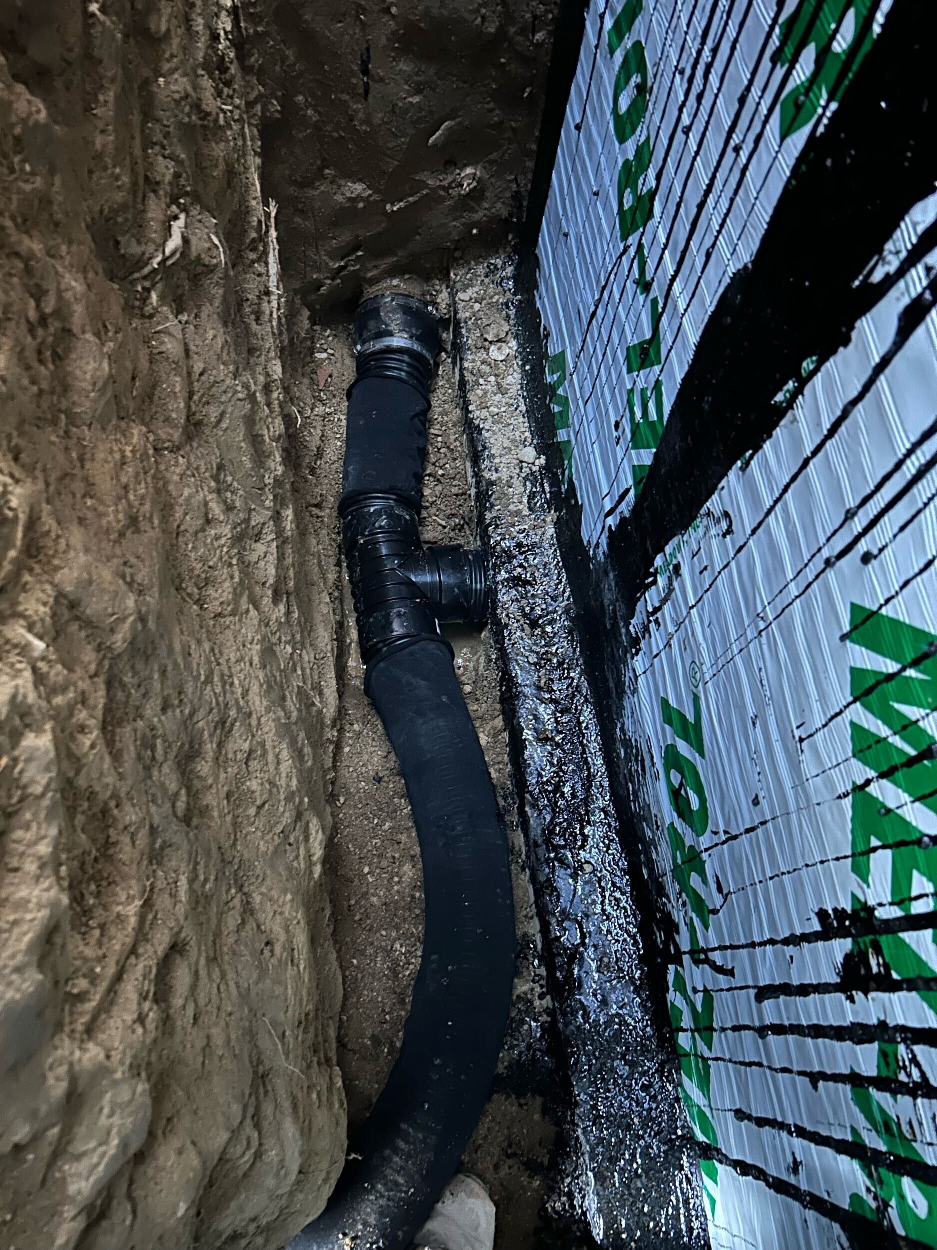 Black drainage pipe running through a narrow dirt trench beside a wrapped wall panel