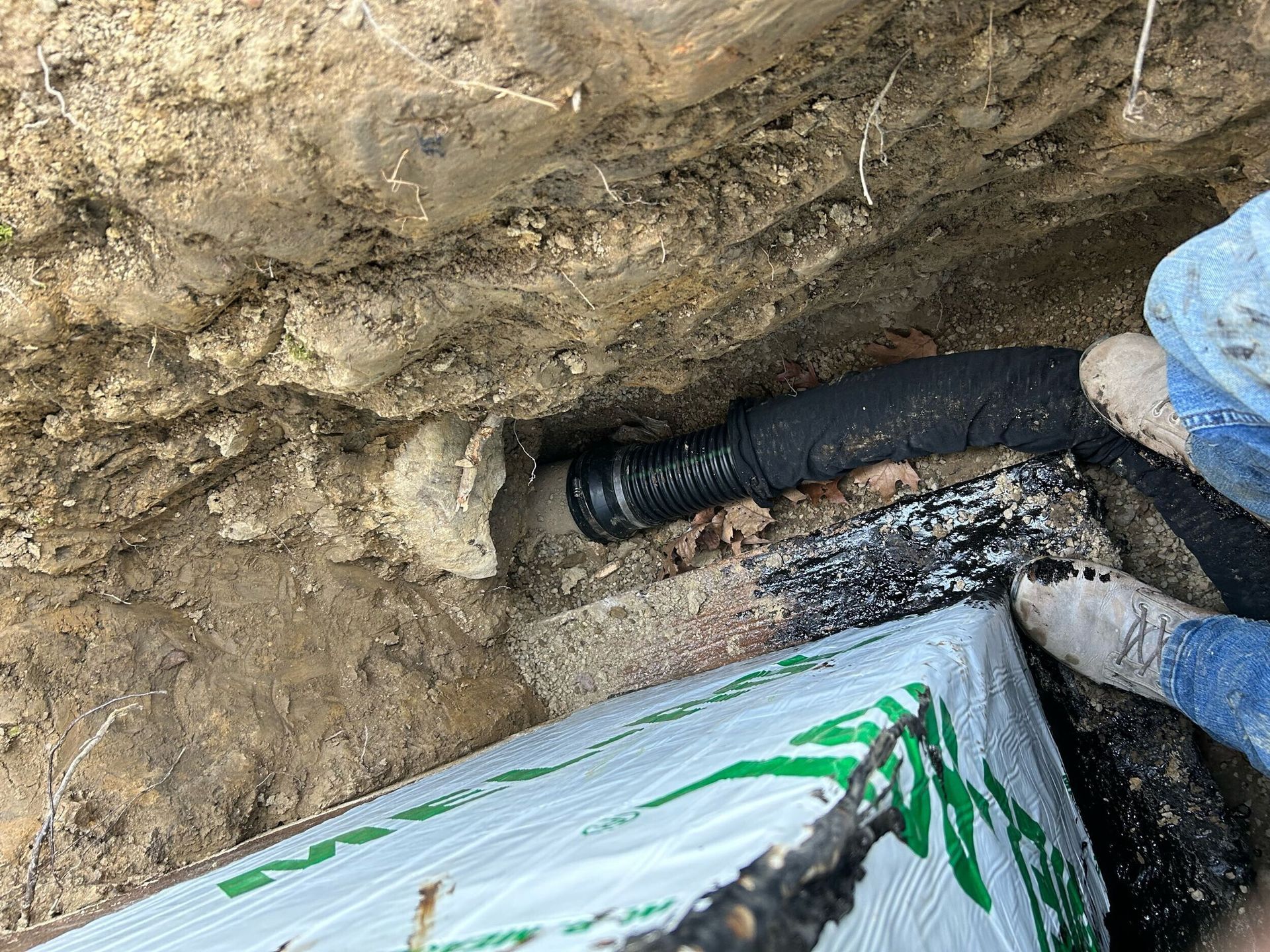 Worker installing black pipe in a muddy trench near wrapped piping and soil wall