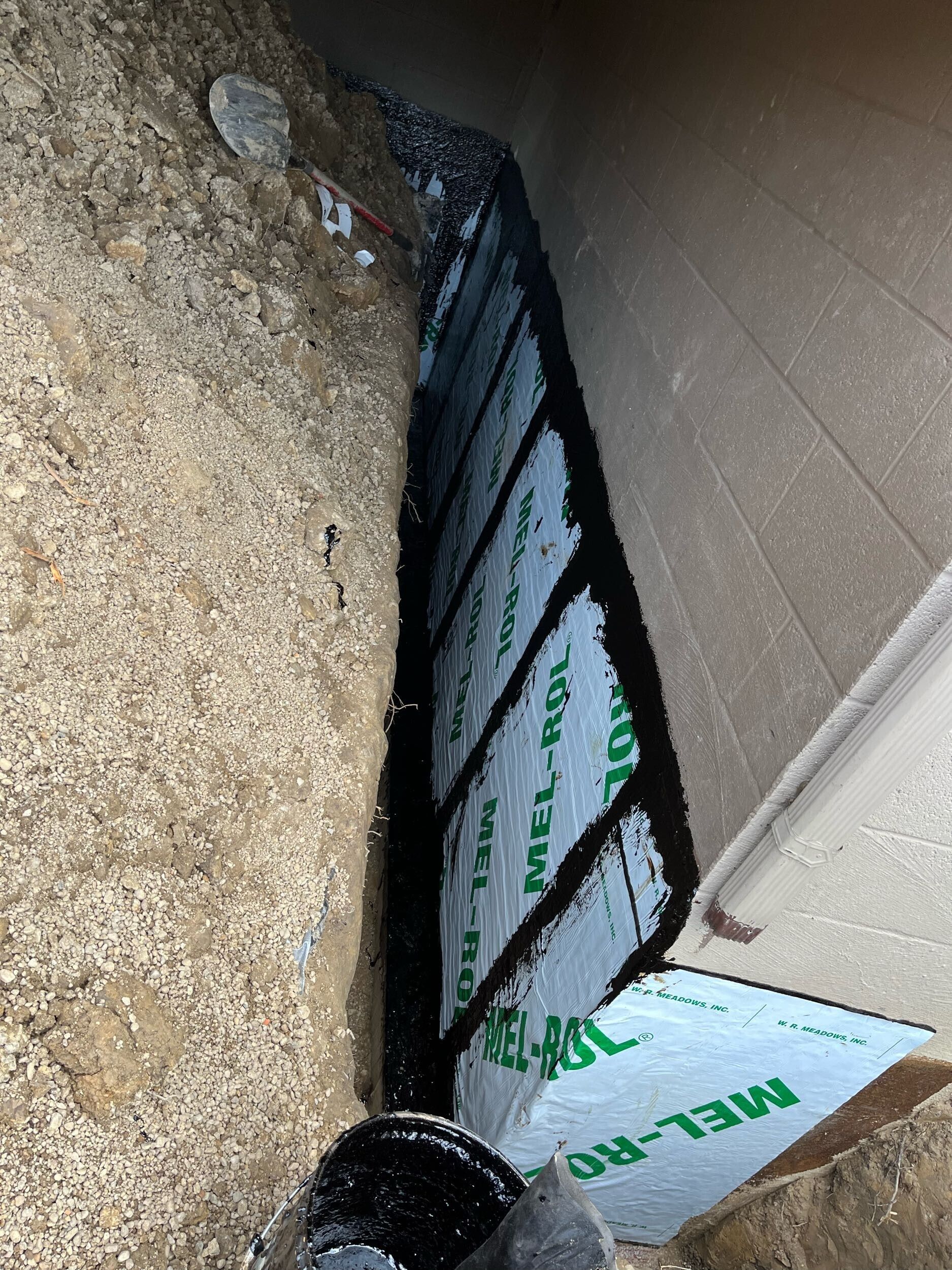 Outdoor basement window well with white insulated panels, black trim, and gravel beside a wall.