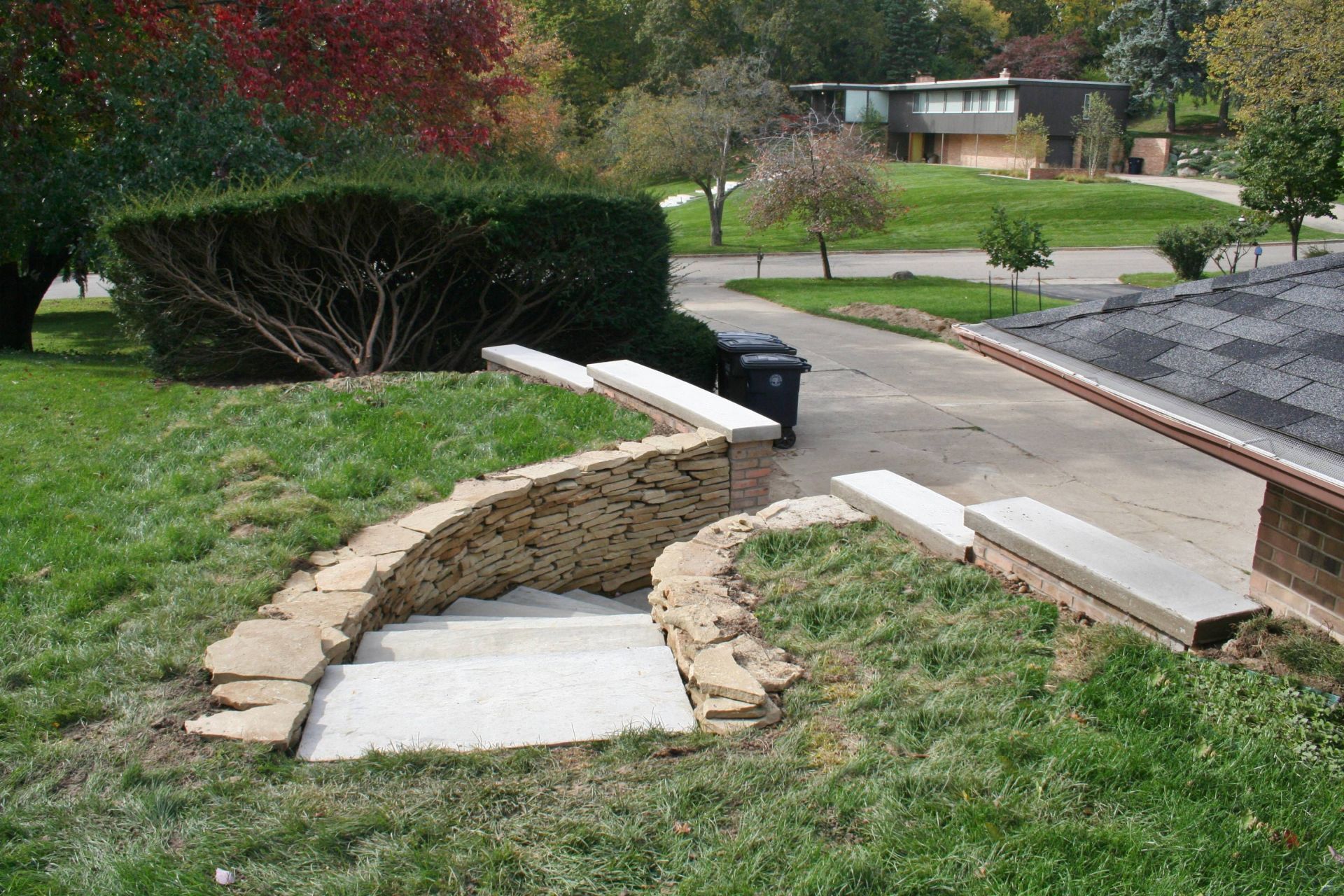 Damaged sidewalk with a collapsed section beside a road and grassy area, near a curb and retaining wall