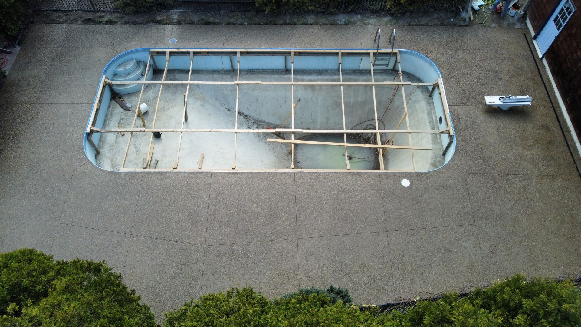 Empty outdoor swimming pool surrounded by concrete and greenery, with pool ladder and railings visible