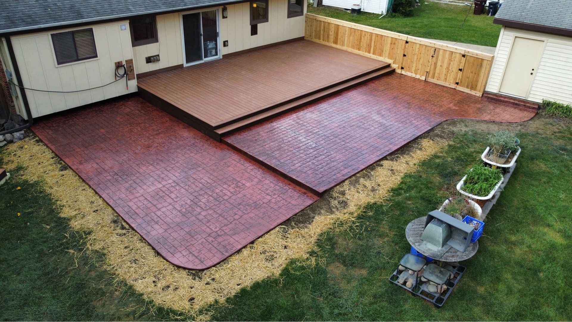 Backyard patio with red and tan paving stones beside a house and lawn, with wheelbarrows on the grass.