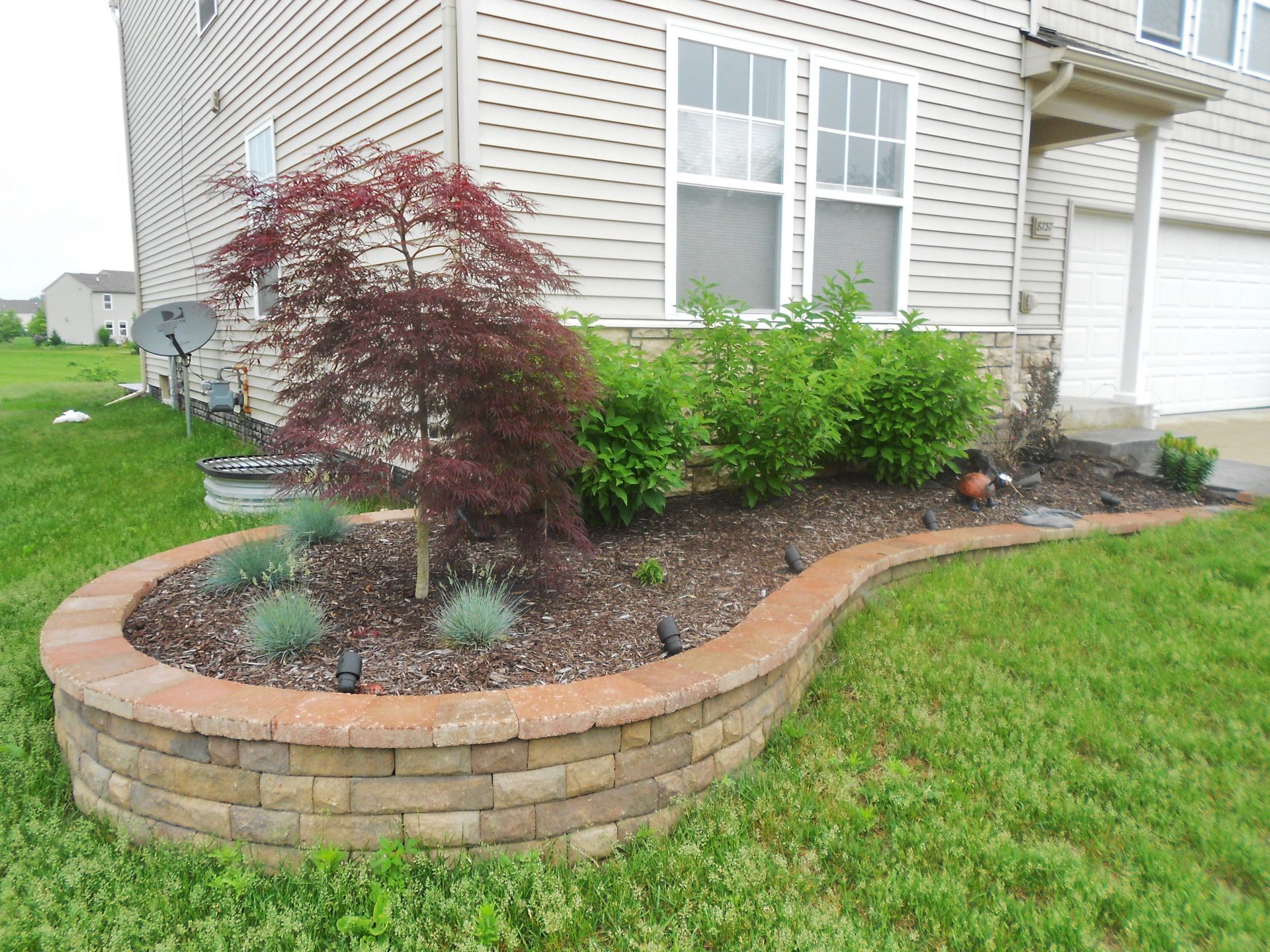 Curved brick flower bed with shrubs and a red-leaf tree beside a house lawn