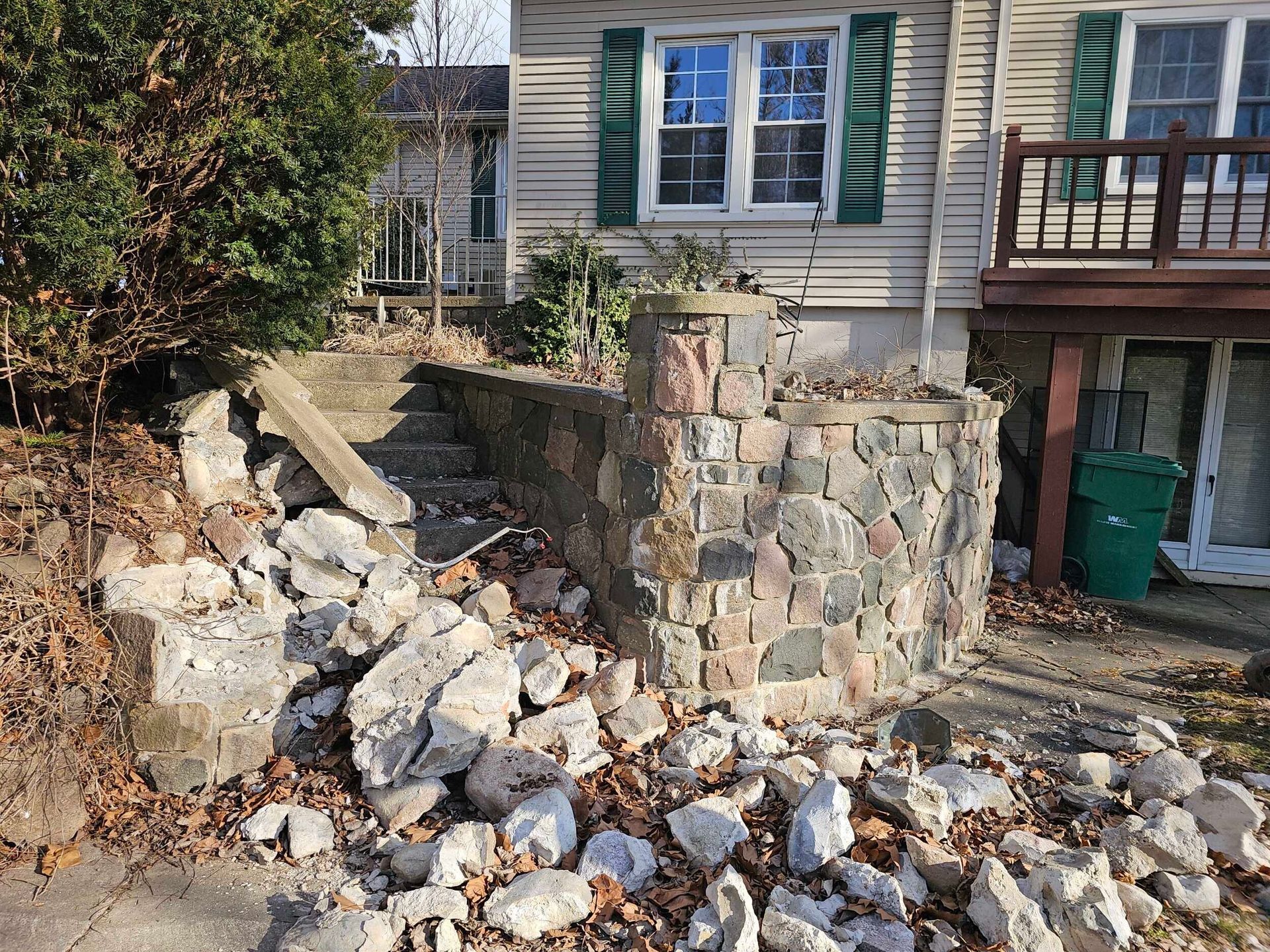 Collapsed stone wall and rubble piled in front of a house with white siding and blue shutters