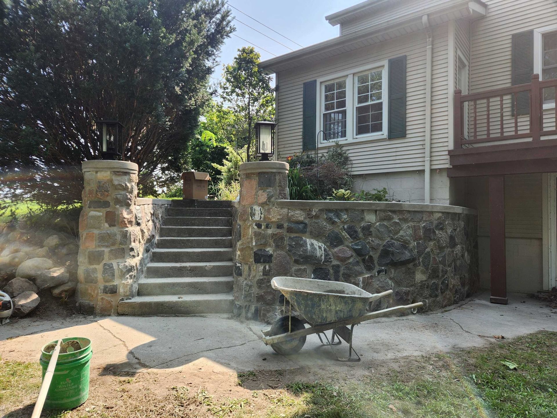 Stone steps and retaining wall beside a house, with a wheelbarrow in the driveway and a green bucket in front.