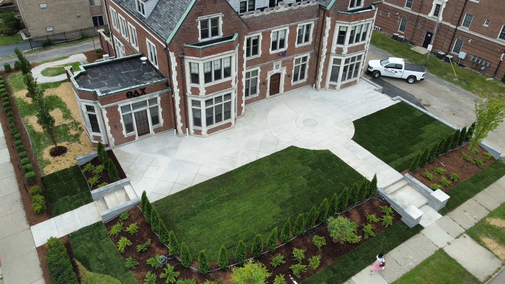 Aerial view of a large brick house with a paved driveway, manicured lawn, and garden beds.