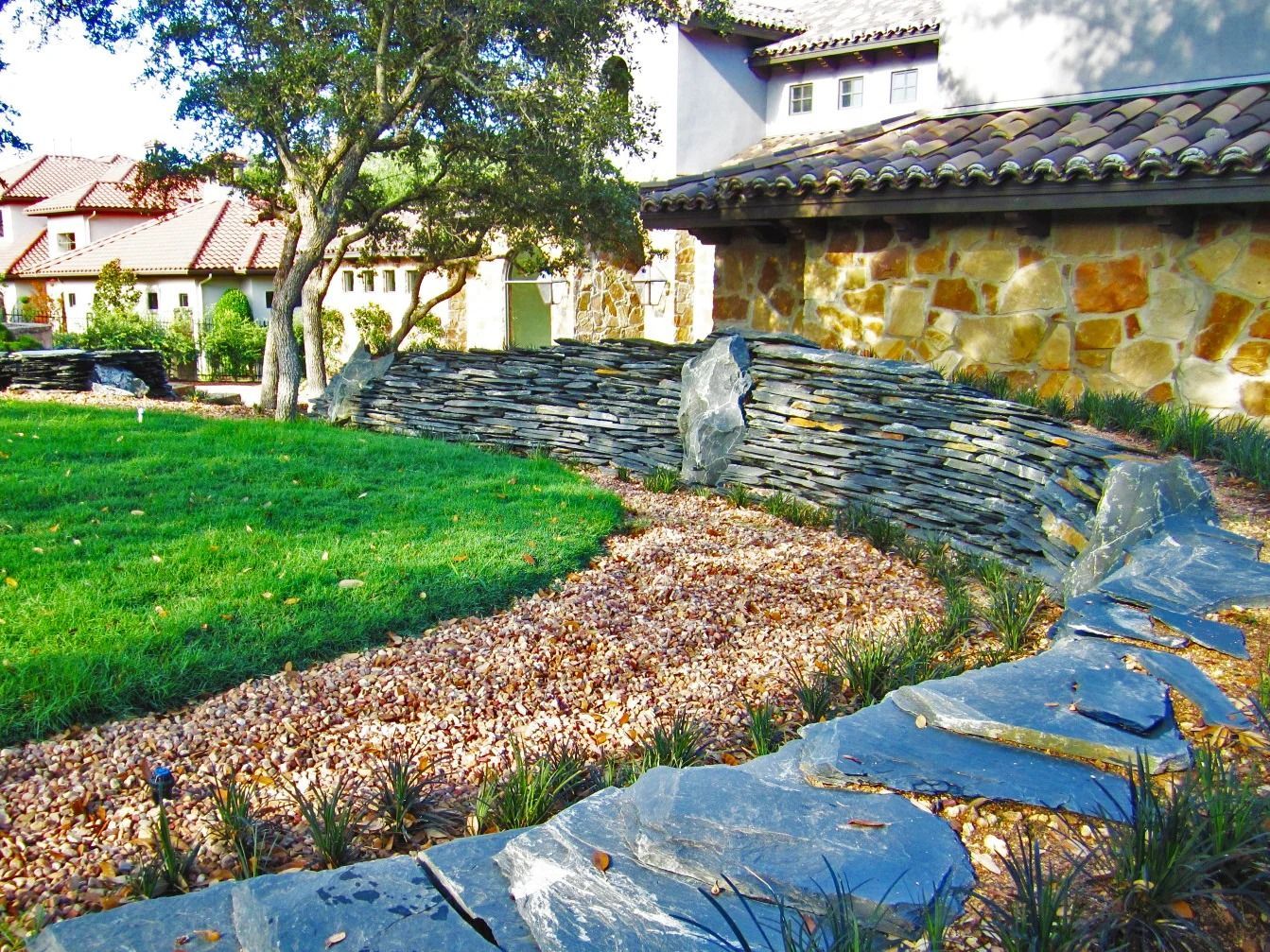 A landscaped yard features a curved slate stone wall, a gravel pathway, and a green lawn in front of a stone building.