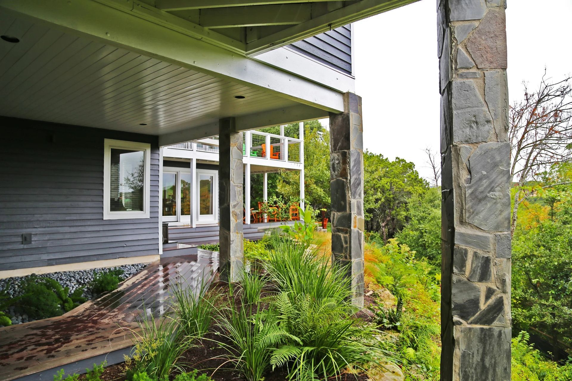 A covered patio with gray siding and stone pillars overlooking a lush, green hillside on a cloudy day.