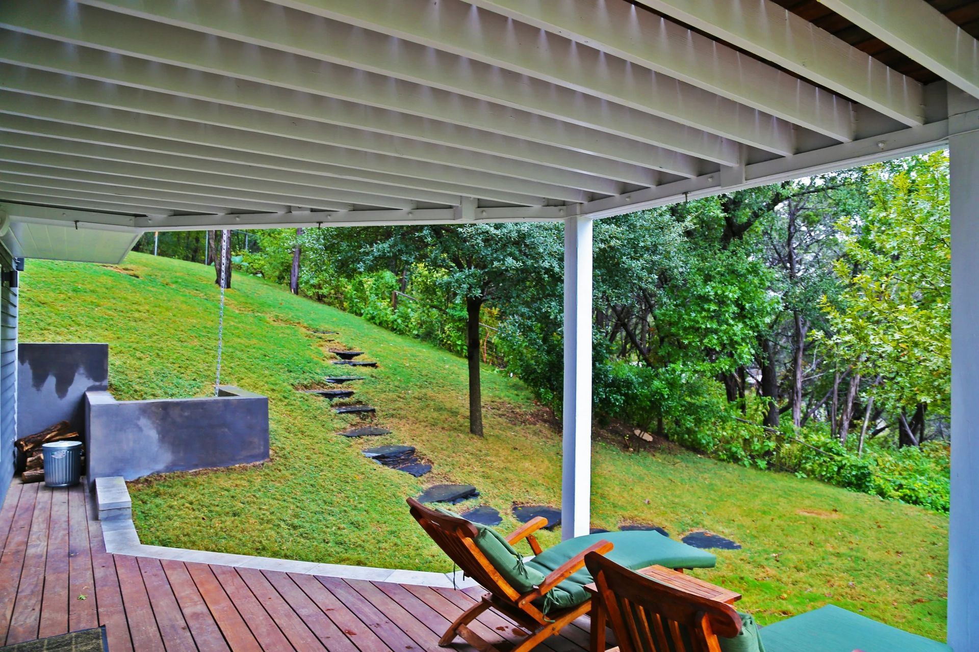 A covered wooden deck with lounge chairs overlooks a grassy, tree-lined hillside with a stone stepping-stone path.