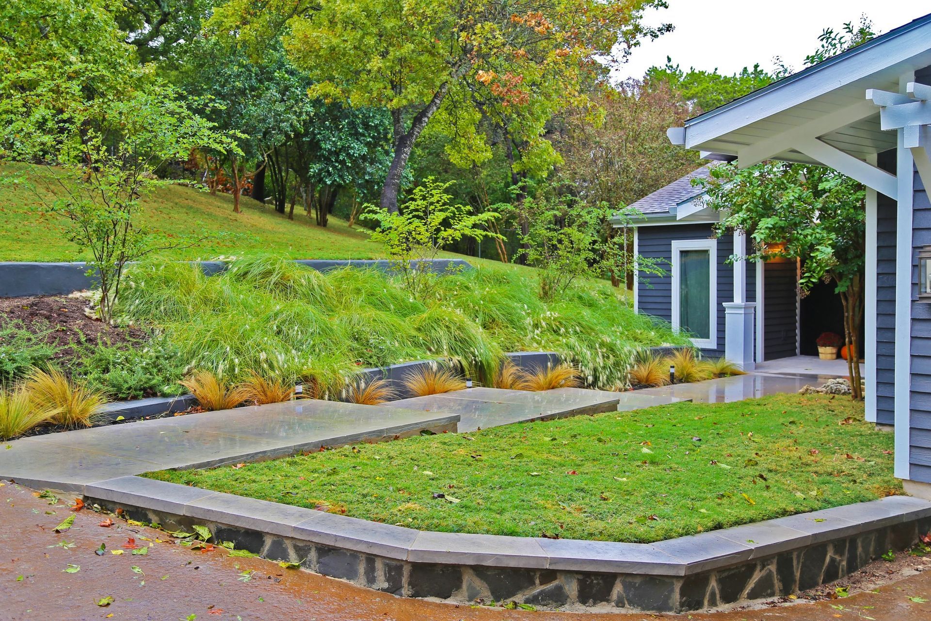A blue-sided cottage sits near a grassy courtyard, framed by a sloped garden, retaining walls, and surrounding trees.