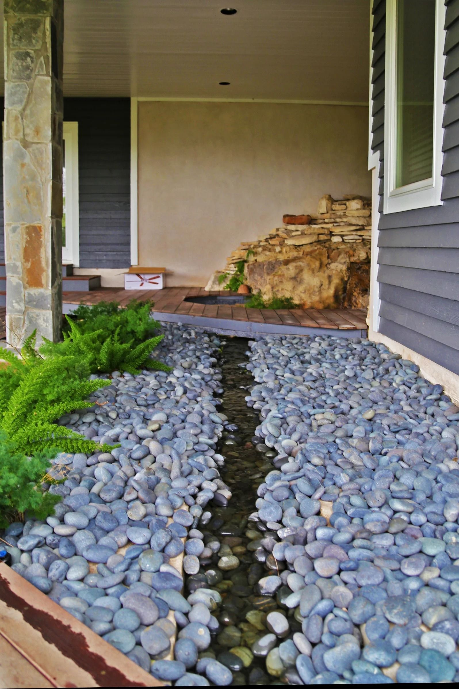A stone-lined water feature flows between gray decorative rocks in a front entryway near a house wall and green plants.