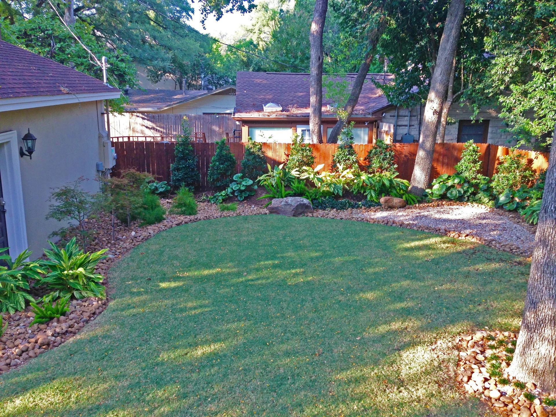 A neatly manicured backyard with a circular green lawn, stone garden beds, and a wooden privacy fence under leafy trees.
