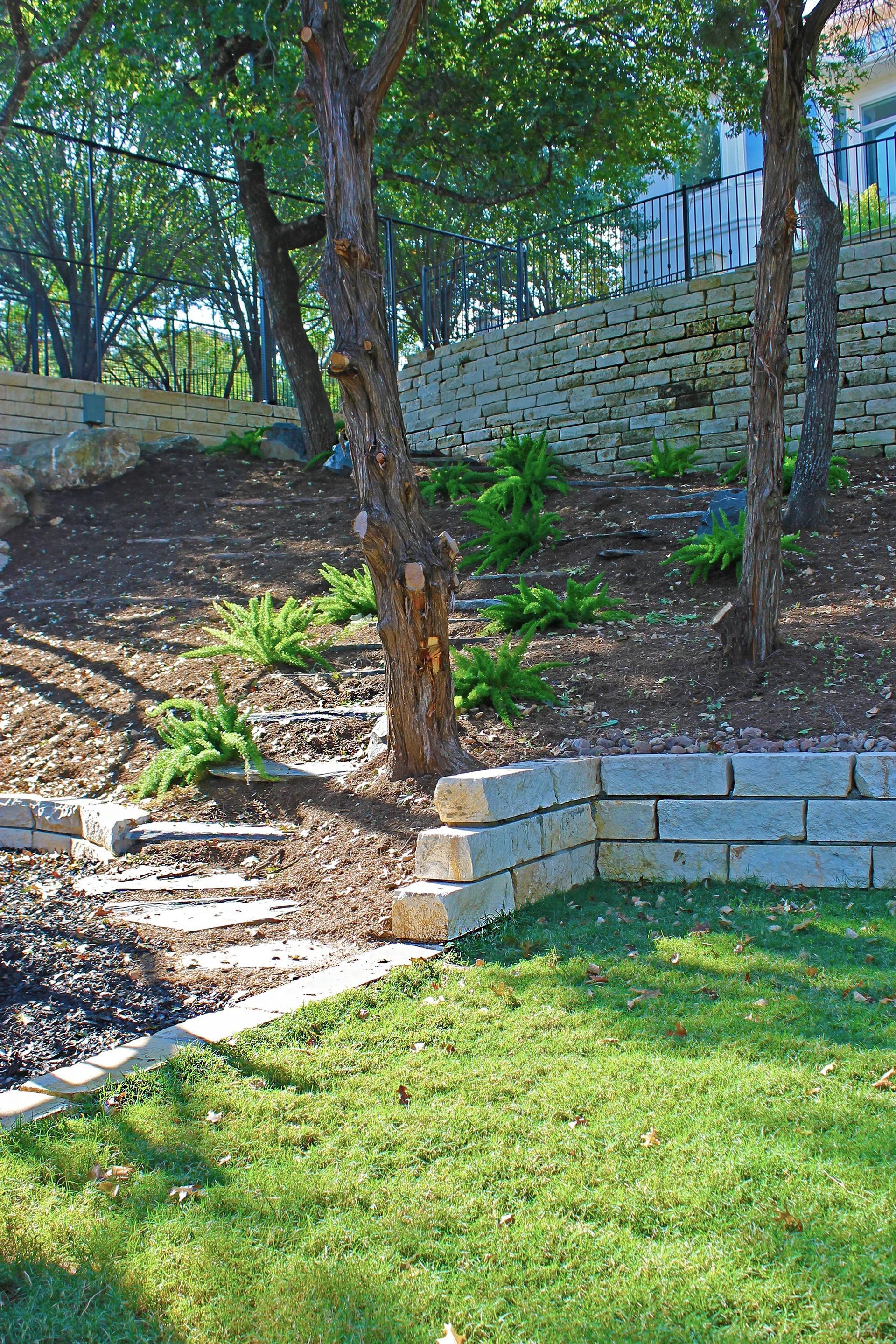 A tiered backyard garden with stone retaining walls, mulched slopes, ferns, and mature trees overlooking a grassy lawn.