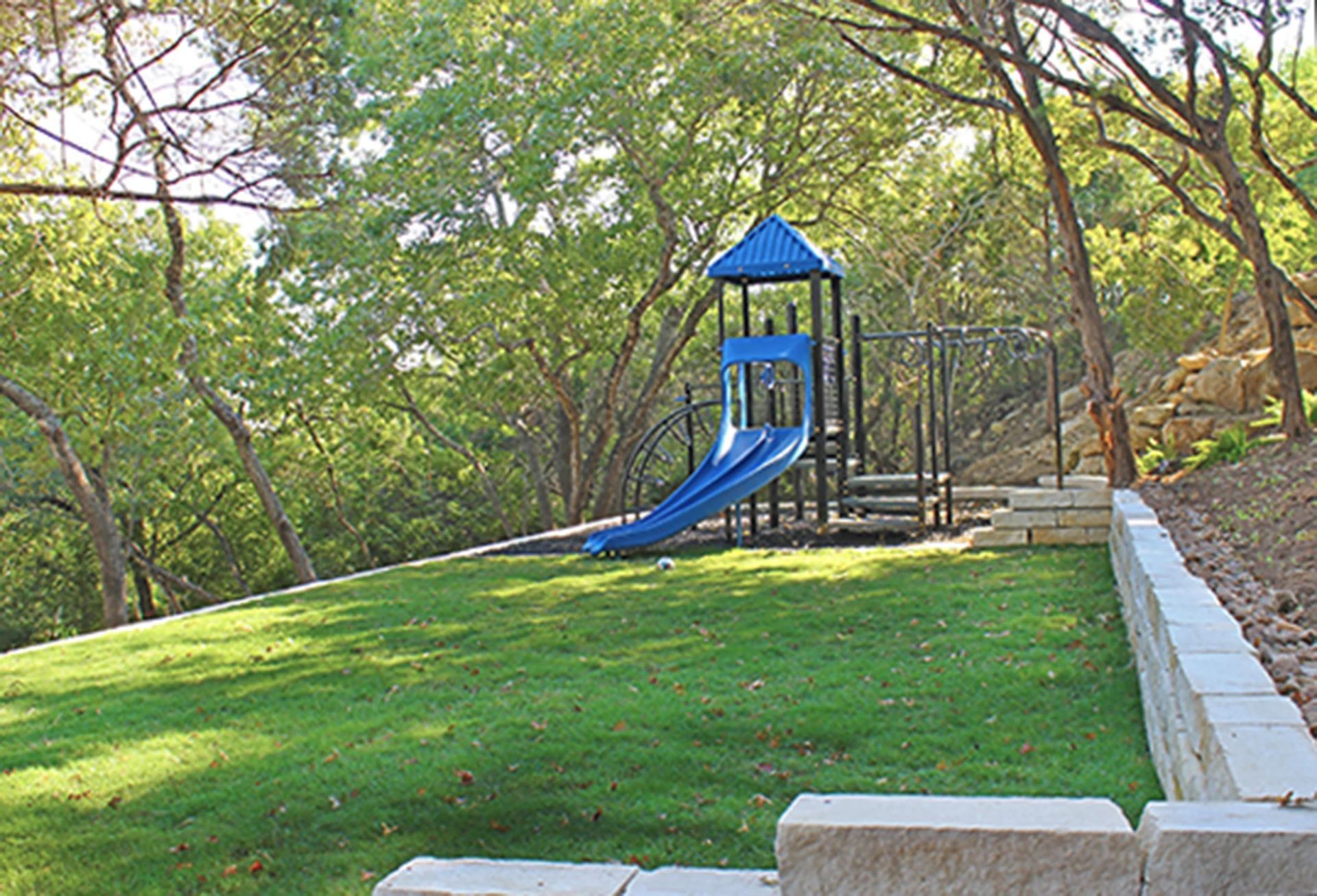 A blue playground set with a slide sits on a grassy lawn next to a low stone retaining wall surrounded by trees.