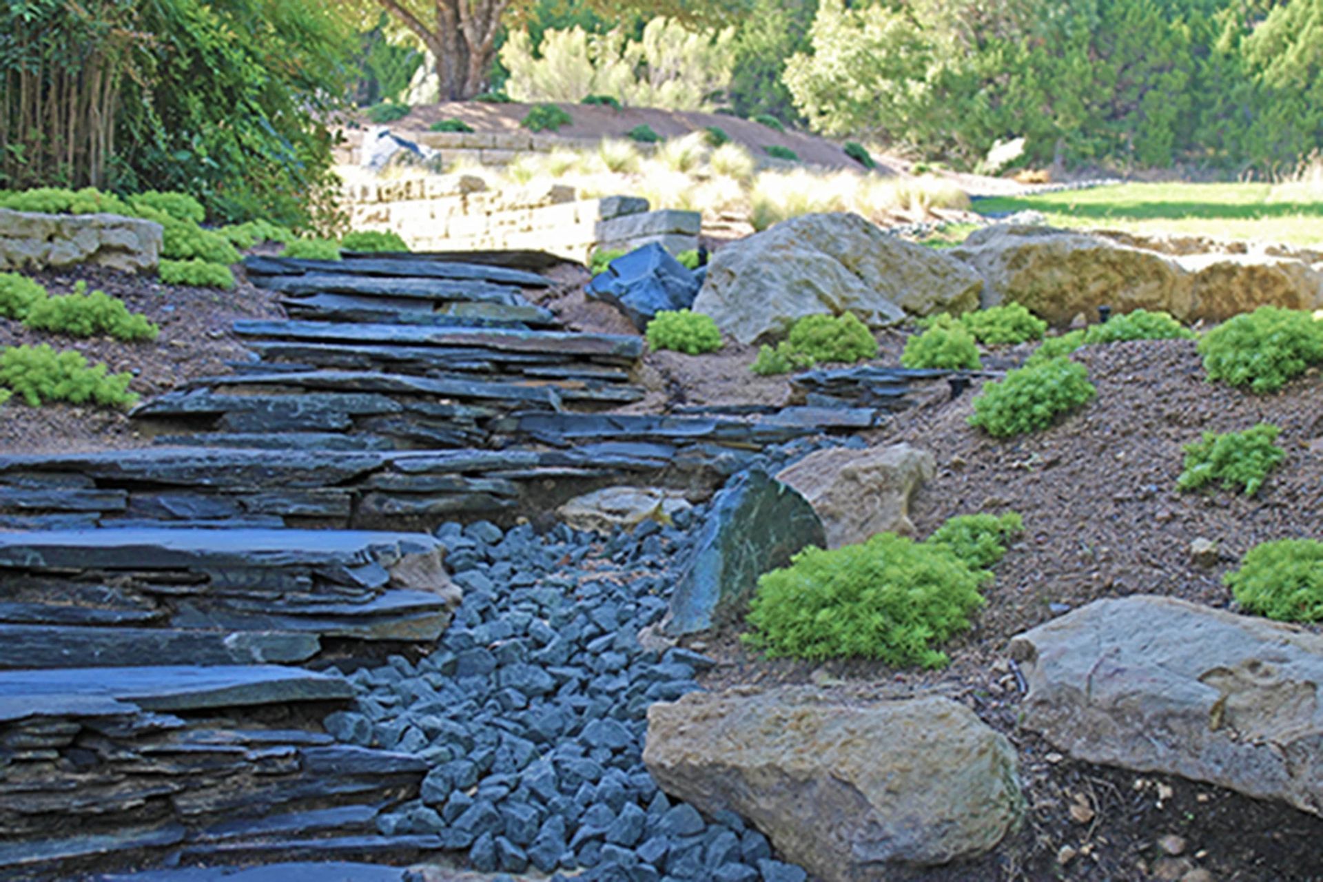 A dry creek bed of dark, layered slate and blue gravel flows through a garden with large rocks and bright green shrubs.