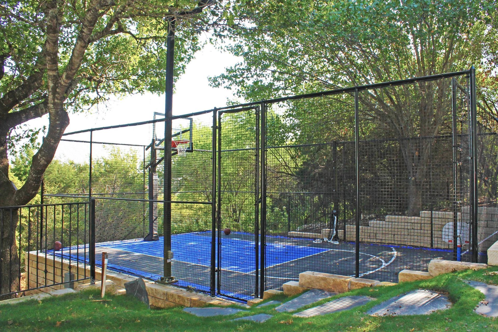A basketball court with a blue and black surface surrounded by a tall metal fence in a lush, wooded outdoor setting.