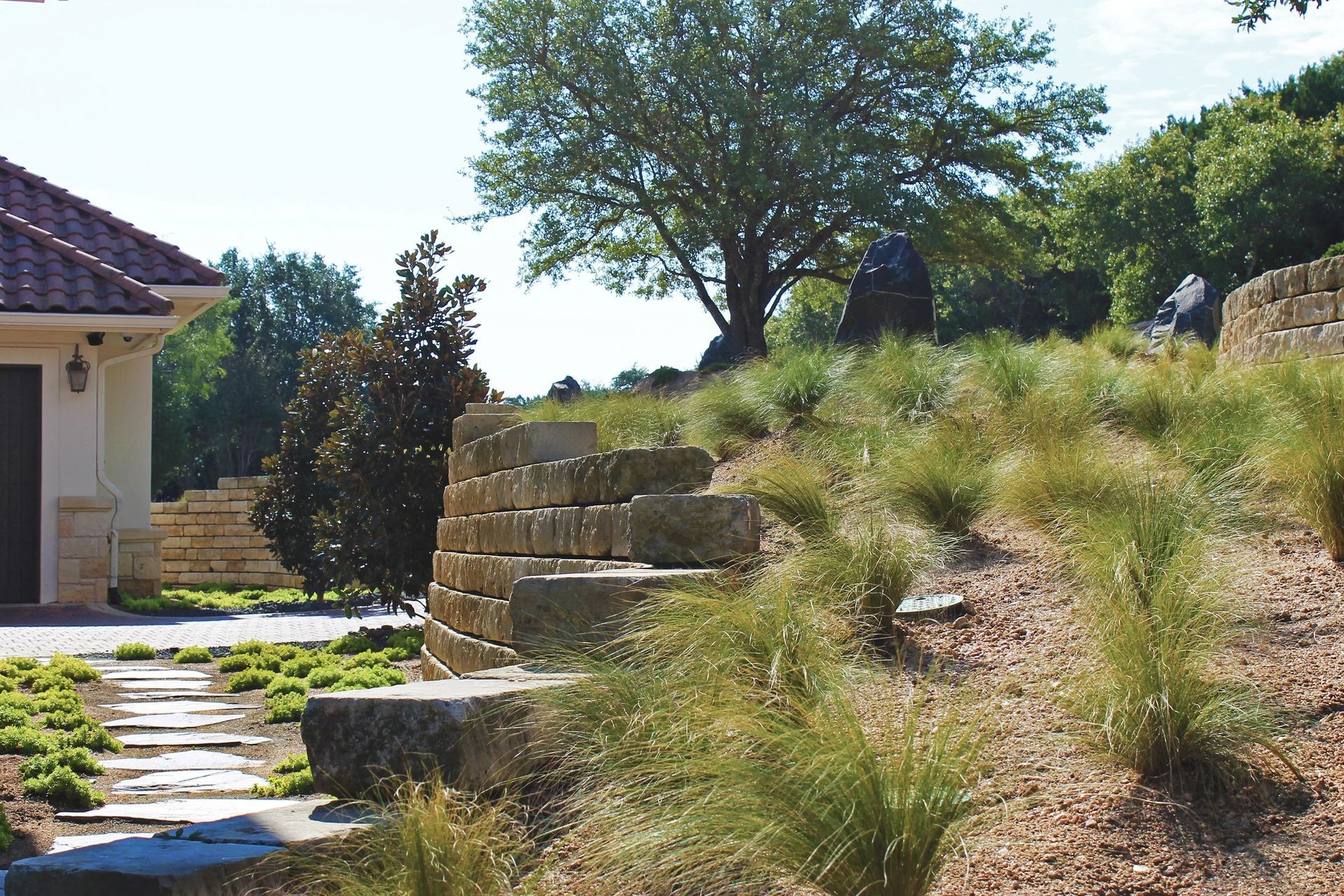 A stone retaining wall sits alongside a garden path near a house, with a grassy hillside and trees in the background.