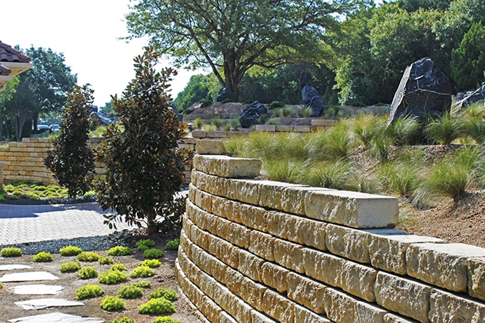 A curved stone retaining wall borders a garden landscape with trees, ornamental grasses, and a stone pathway.