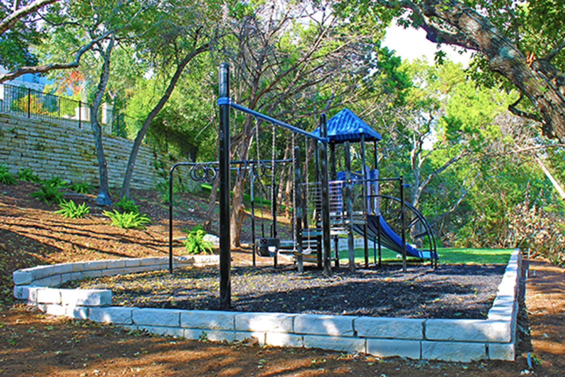 A playground with a blue slide, climbing structure, and swings, surrounded by a stone retaining wall in a wooded area.