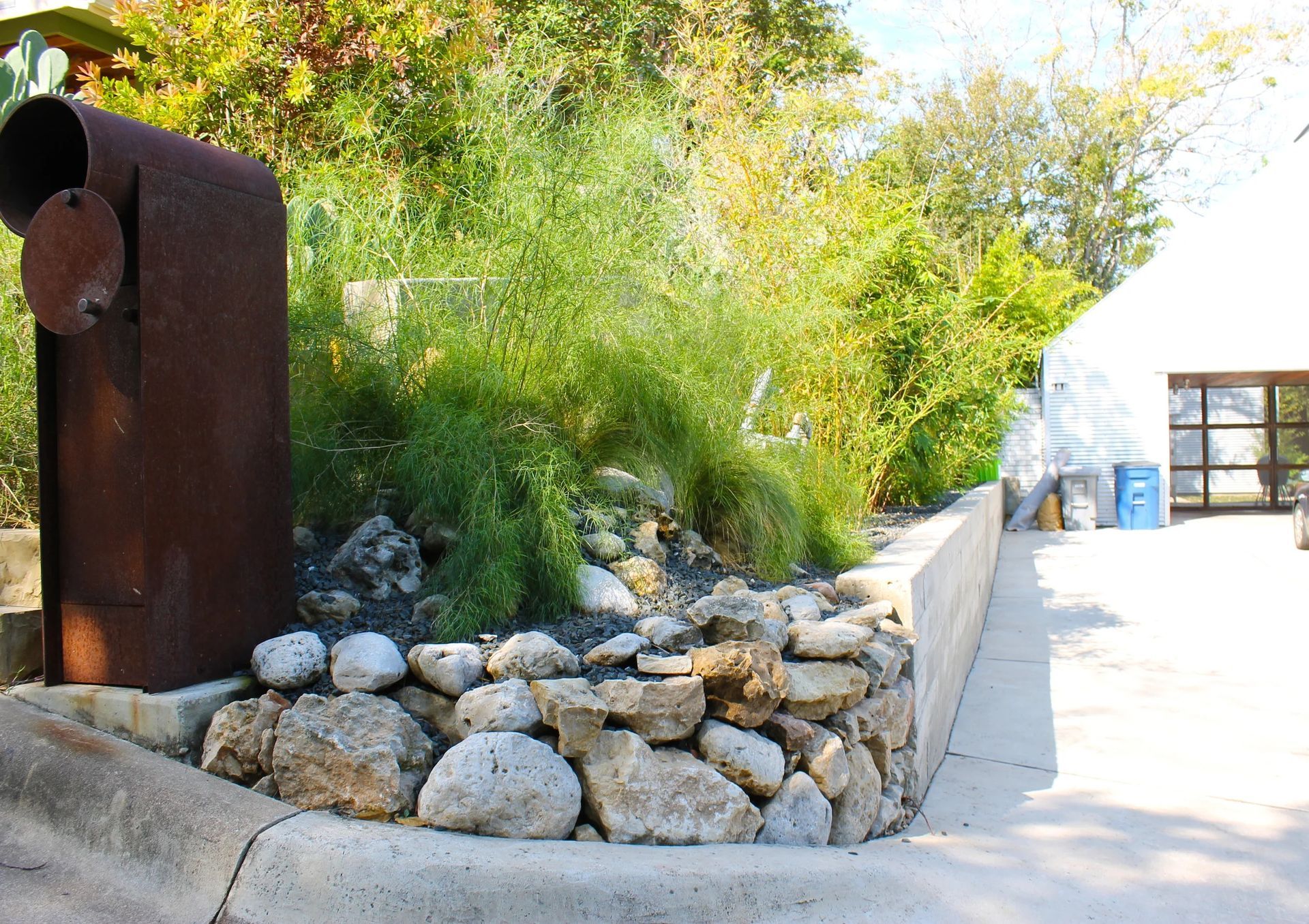 A rusted mailbox stands next to a rock retaining wall and green plants along a paved driveway.