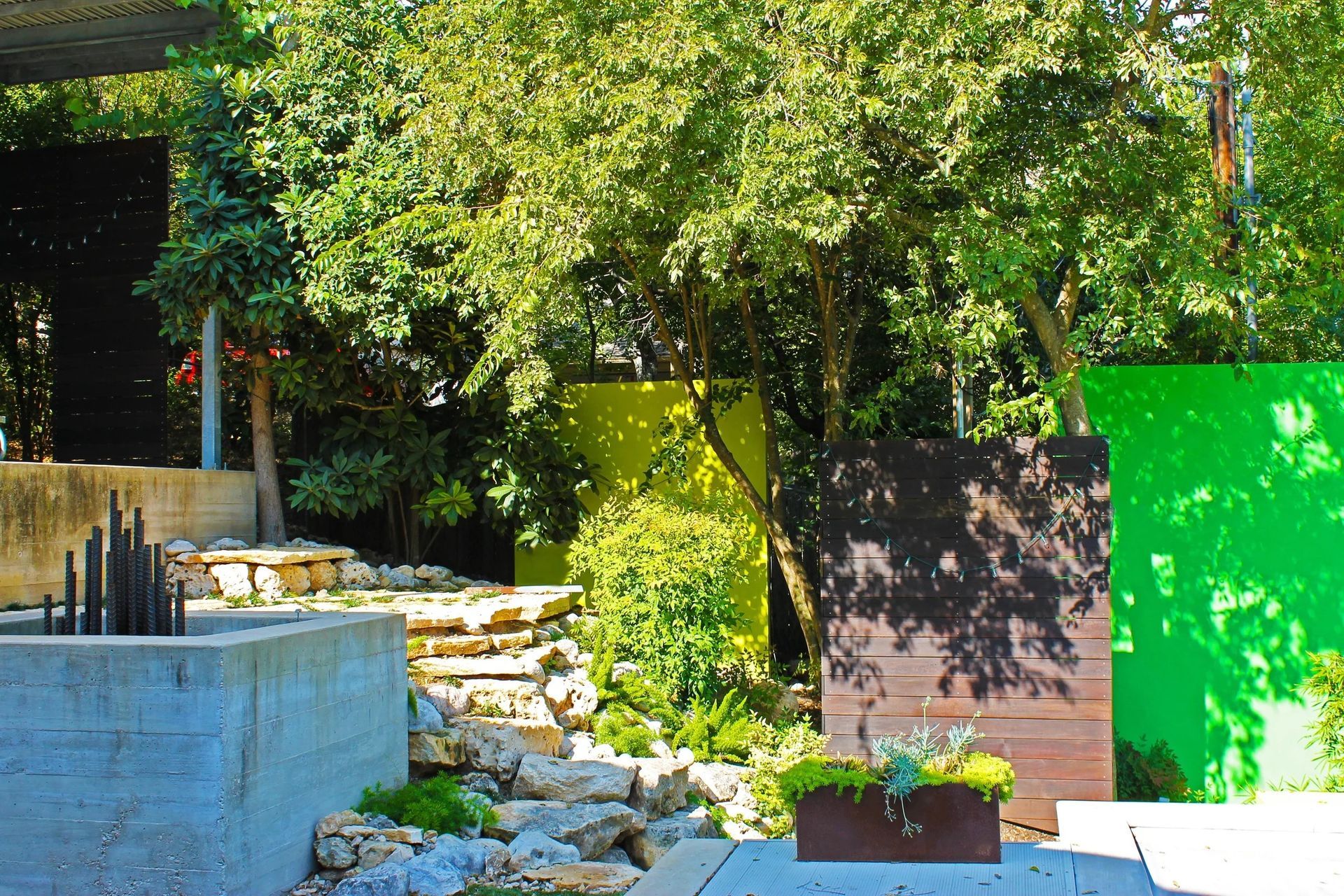 A garden landscape featuring a stone planter, textured rock steps, a dark wood panel, and vibrant lime and green backdrops.
