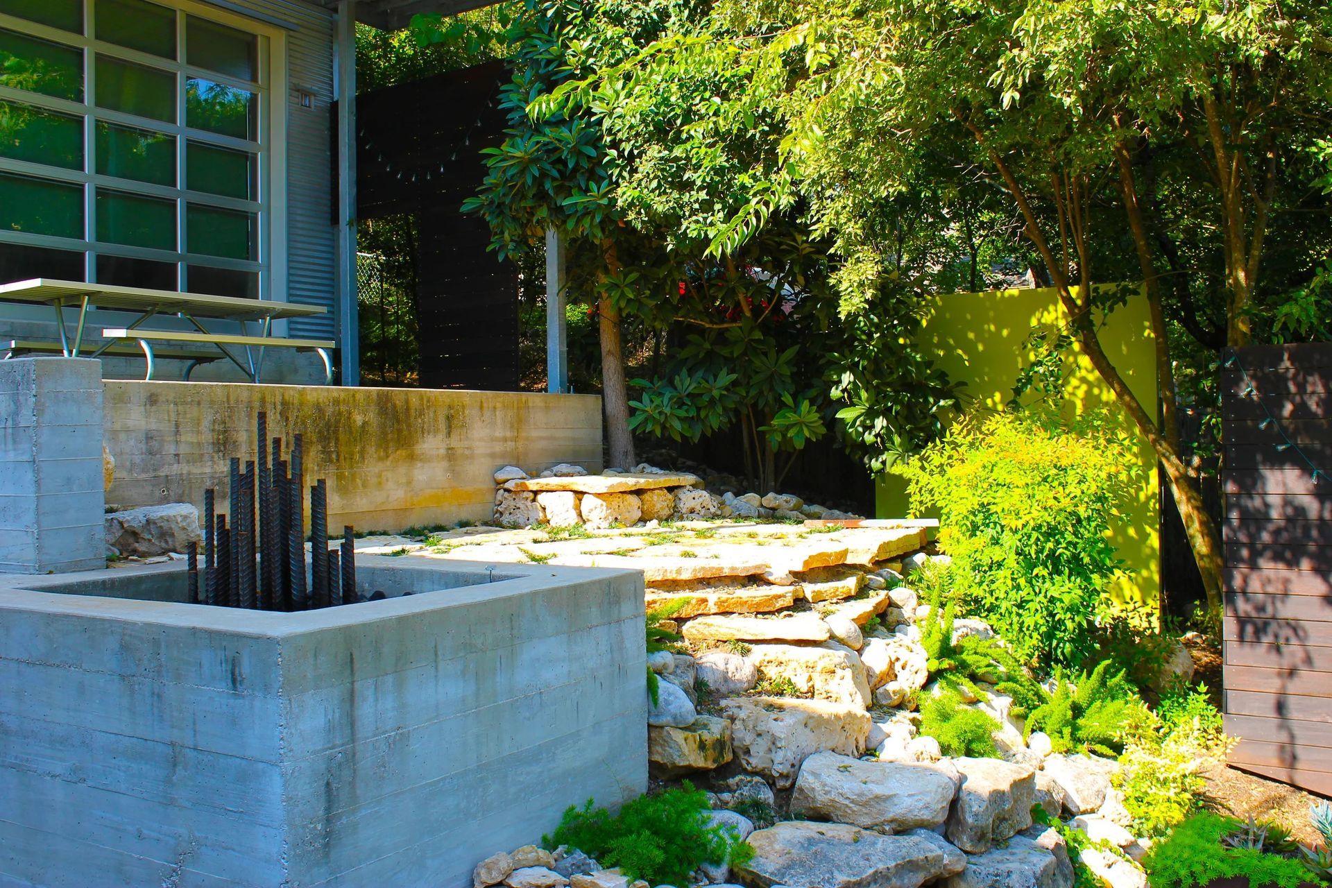 A concrete fire pit sits in a garden next to terraced stone steps leading to a patio with a metal table and chairs.