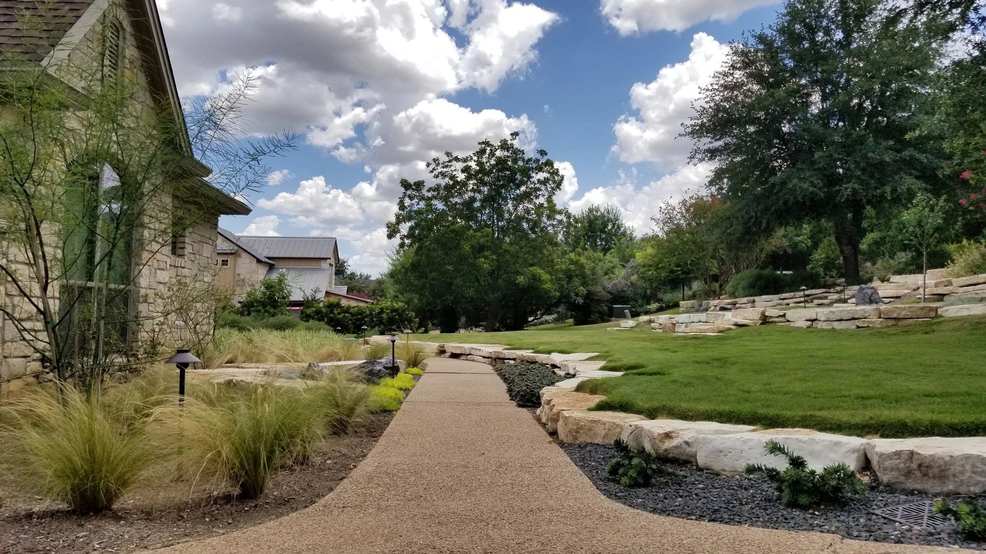 A gravel path leads through a landscaped yard with stone retaining walls, green grass, and trees under a cloudy blue sky.
