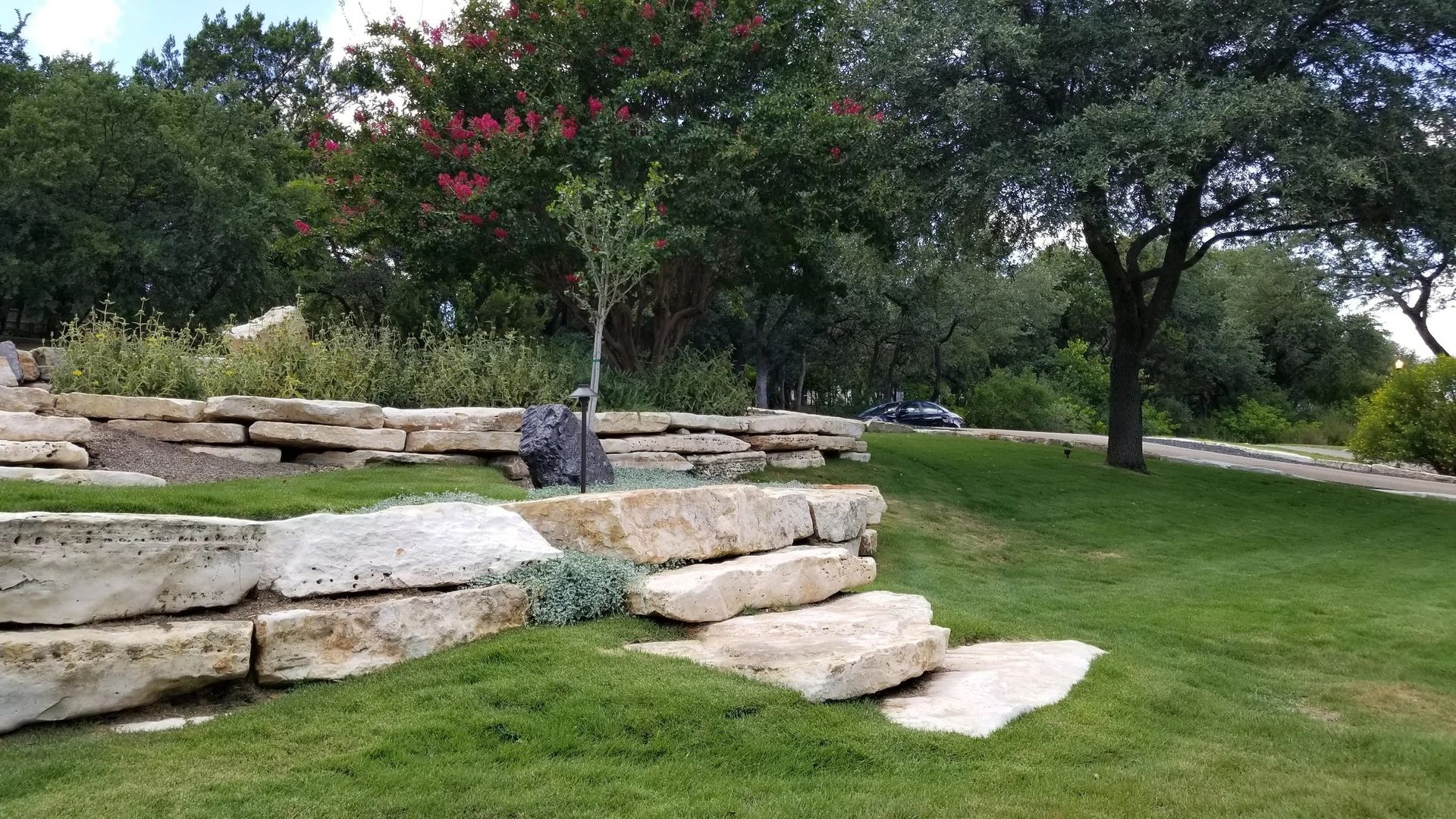 Tiered limestone retaining walls integrated into a landscaped grassy slope with mature trees and a paved path.