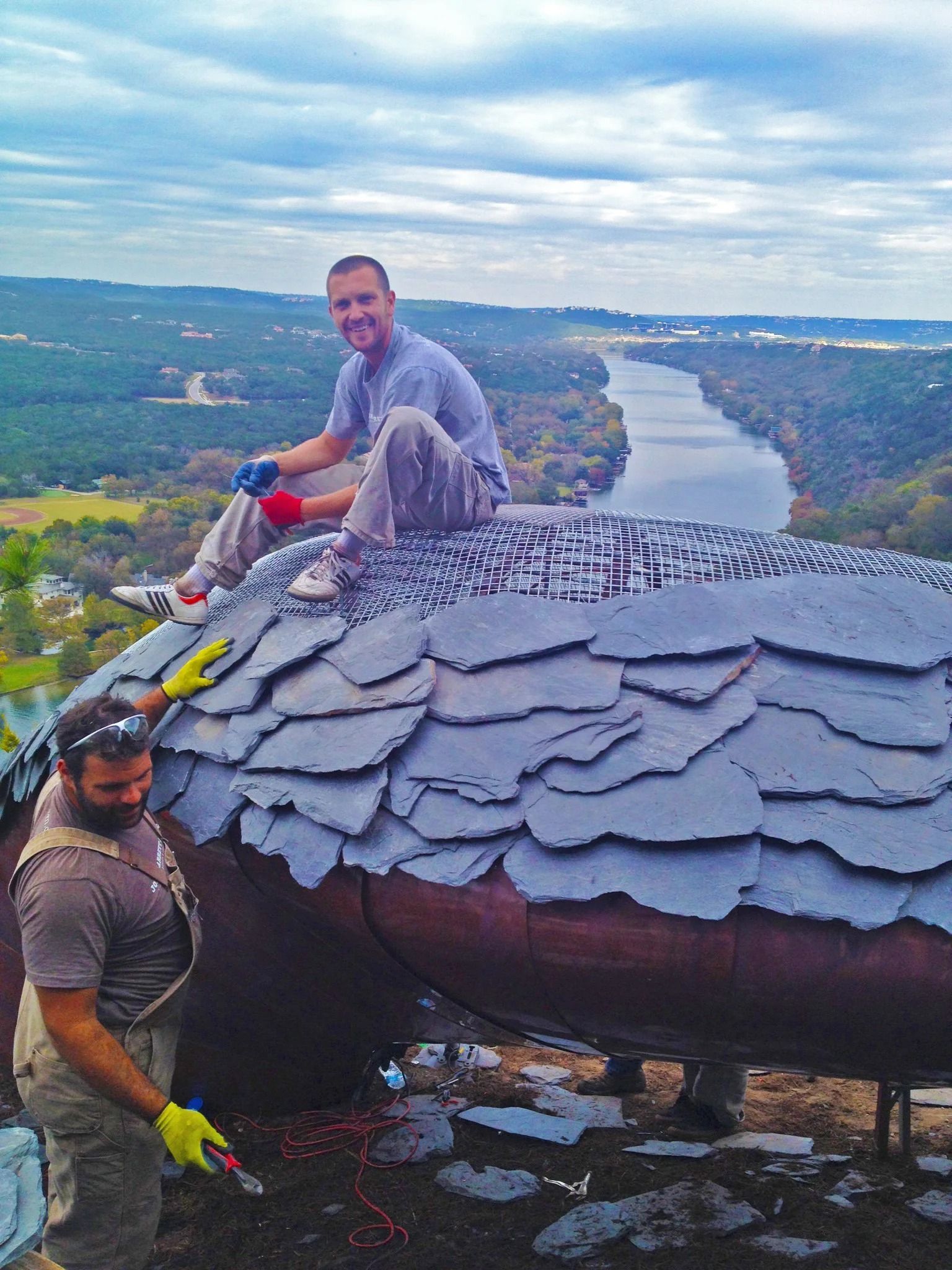 Two people work on a metal structure covered in slate shingles overlooking a river and valley landscape.