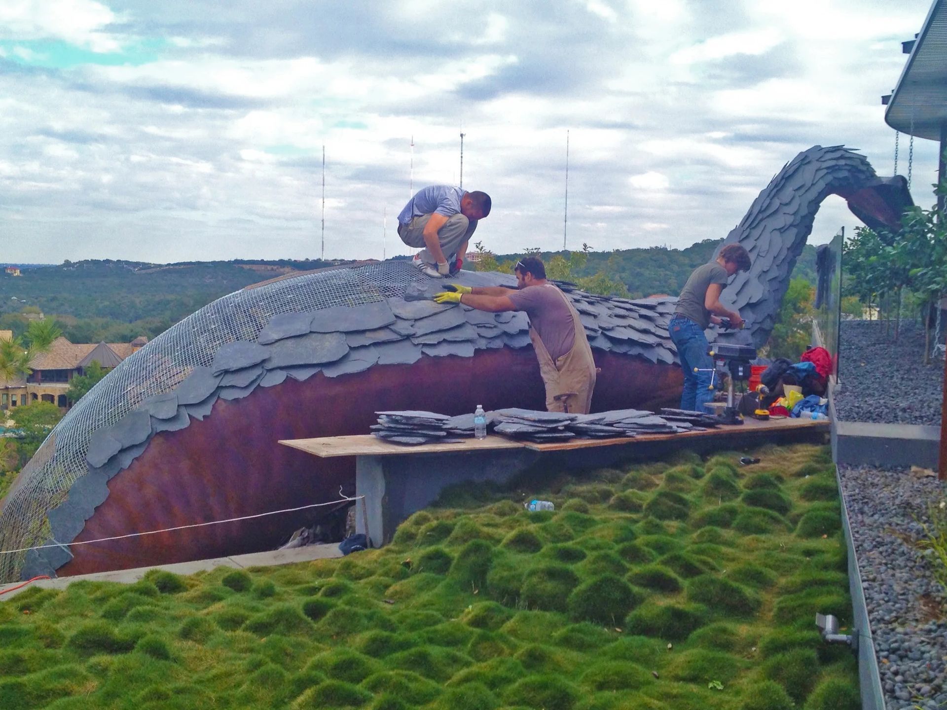 Workers installing slate shingles on a giant dinosaur sculpture roof, outdoors.