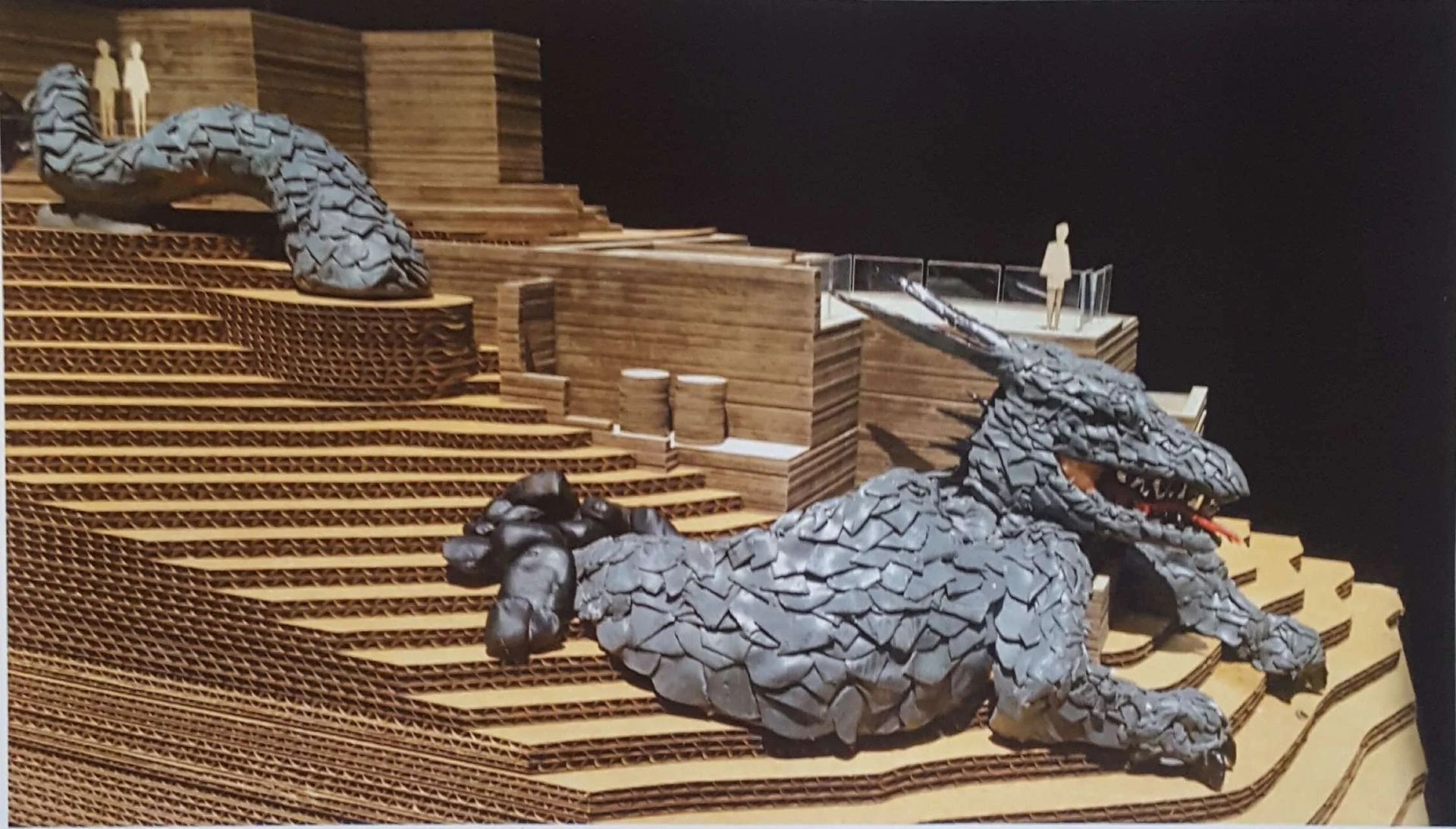 A scale model of a dragon sculpture resting on stepped, wood-textured terraces with small human figures for scale.