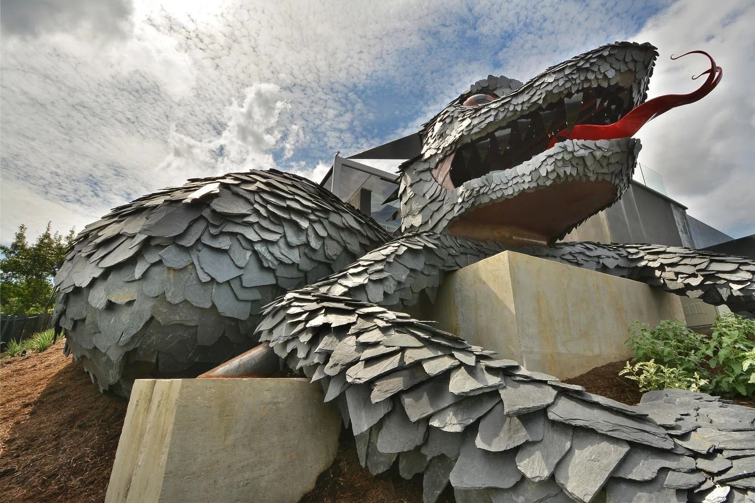 A large dragon sculpture covered in gray slate shingles, featuring a bright red tongue, outdoors against a cloudy sky.