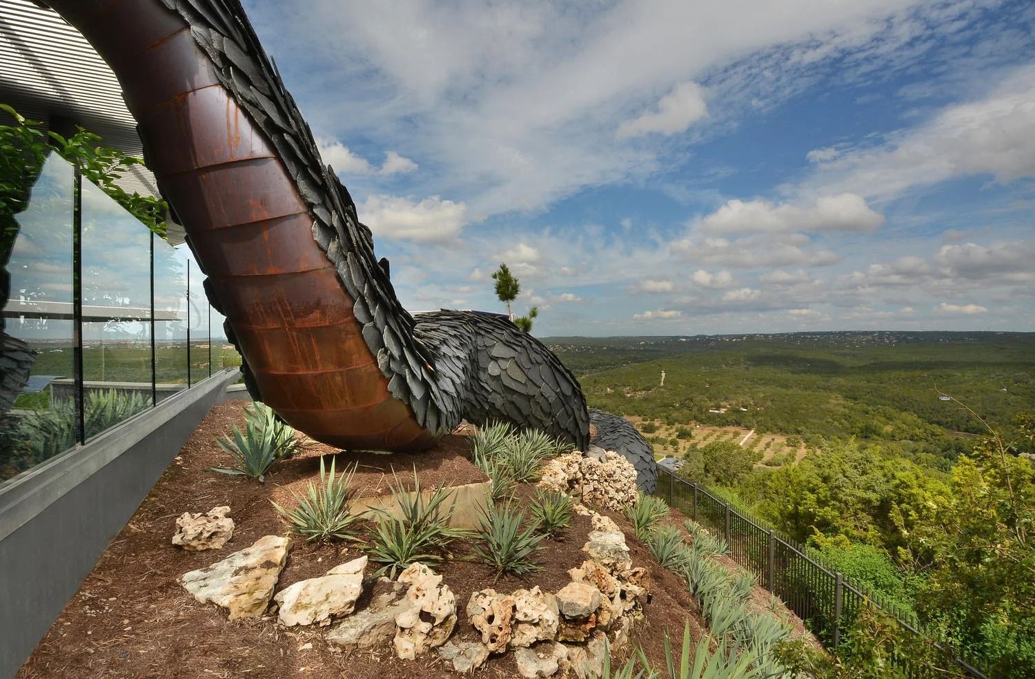 A dark, metallic, winding sculpture sits on a landscaped terrace overlooking a vast, green forest under a cloudy sky.