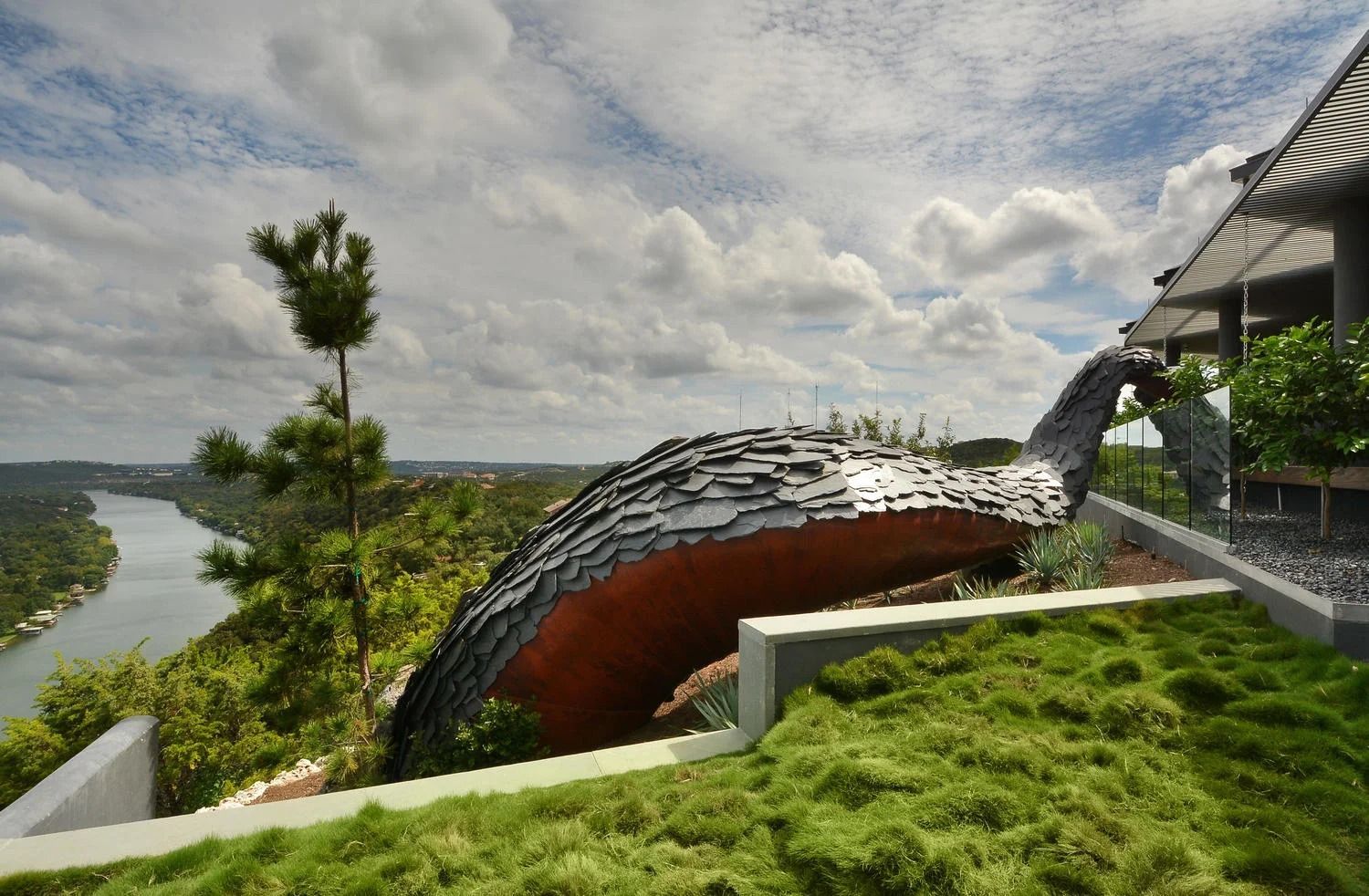 A large, curved sculpture with a metallic, layered surface overlooks a river winding through a forested landscape.