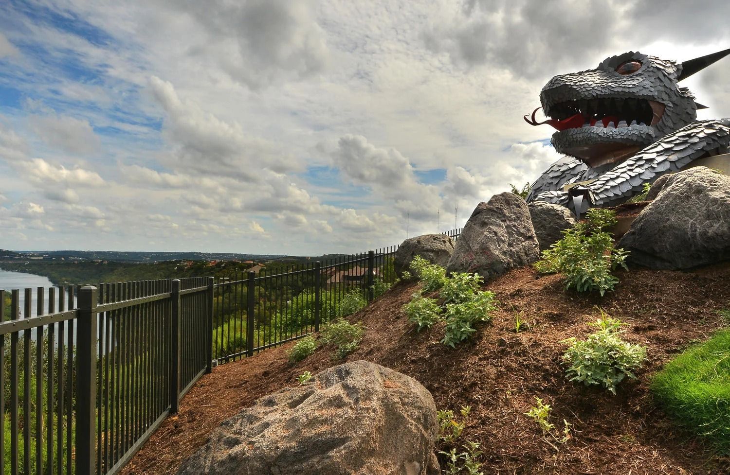 A metal dragon sculpture sits on a rocky hill overlooking a river and distant landscape under a cloudy sky.