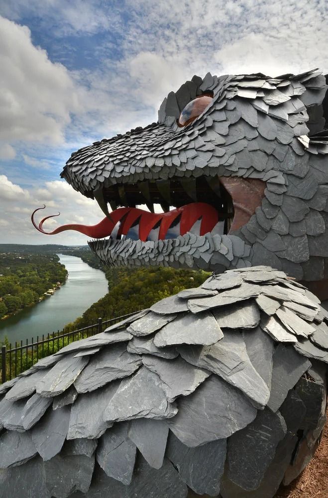 A large, slate-scaled serpent sculpture with a red tongue overlooks a winding river under a cloudy sky.