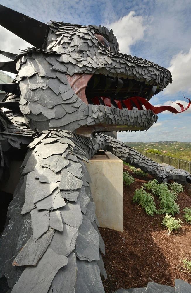 Close-up of a large dragon sculpture made of dark gray stone plates, with a long red tongue, overlooking a grassy valley.