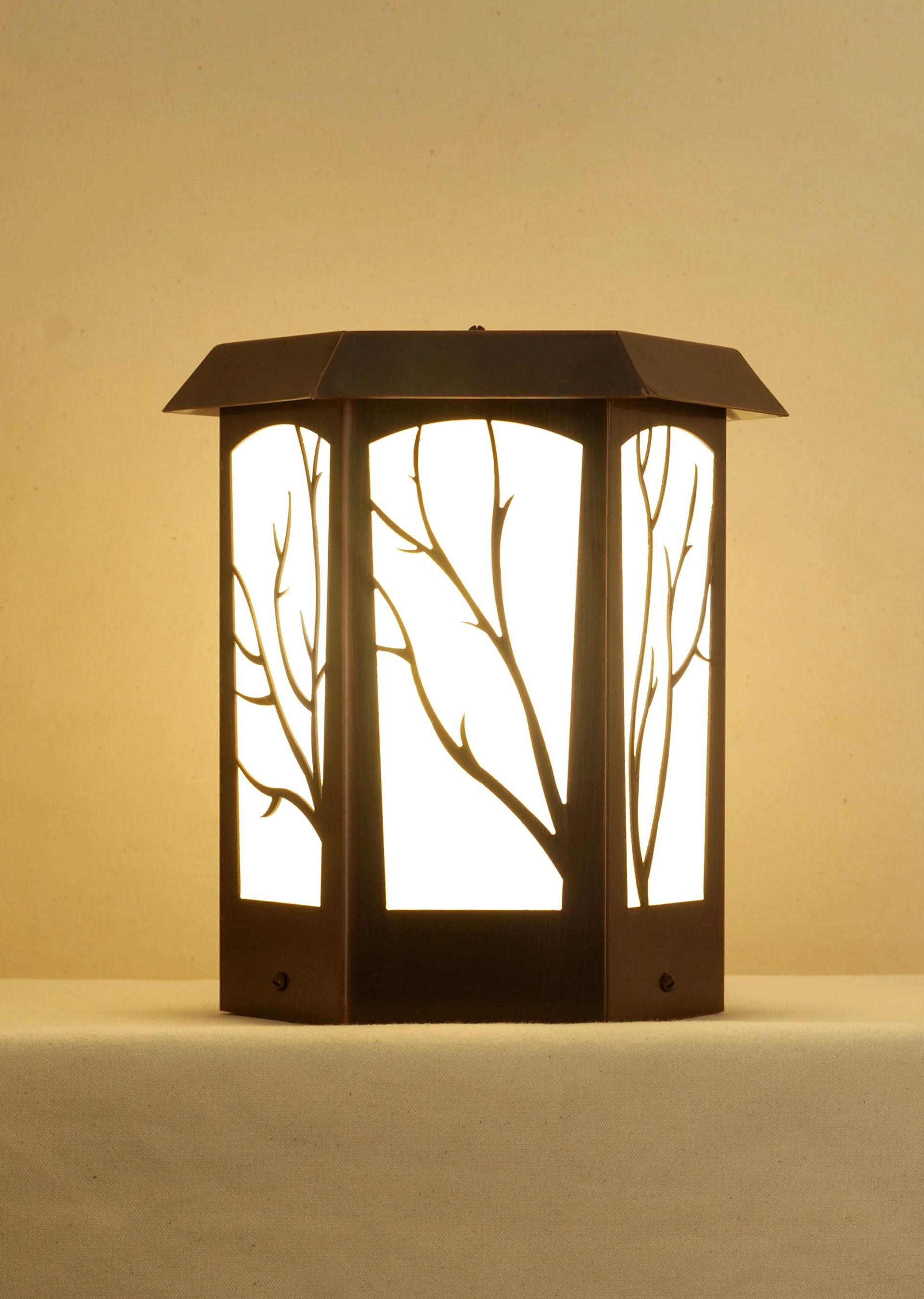 A hexagonal lantern with a dark metal frame and backlit panels featuring a black tree branch silhouette design.
