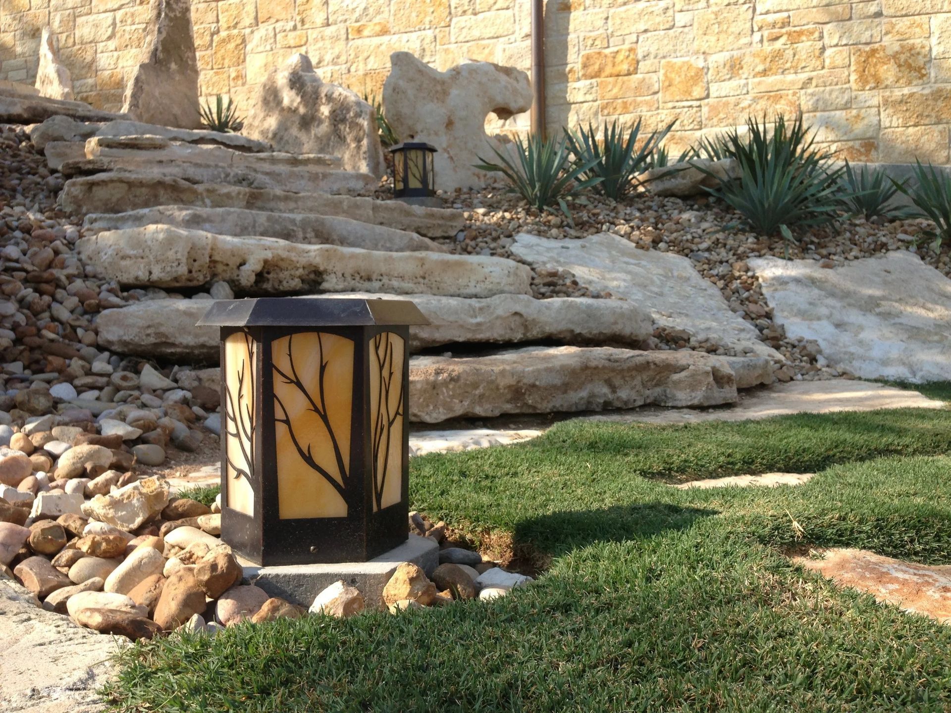 A decorative outdoor lantern sits on stone steps near a small patch of grass, agave plants, and a rock retaining wall.