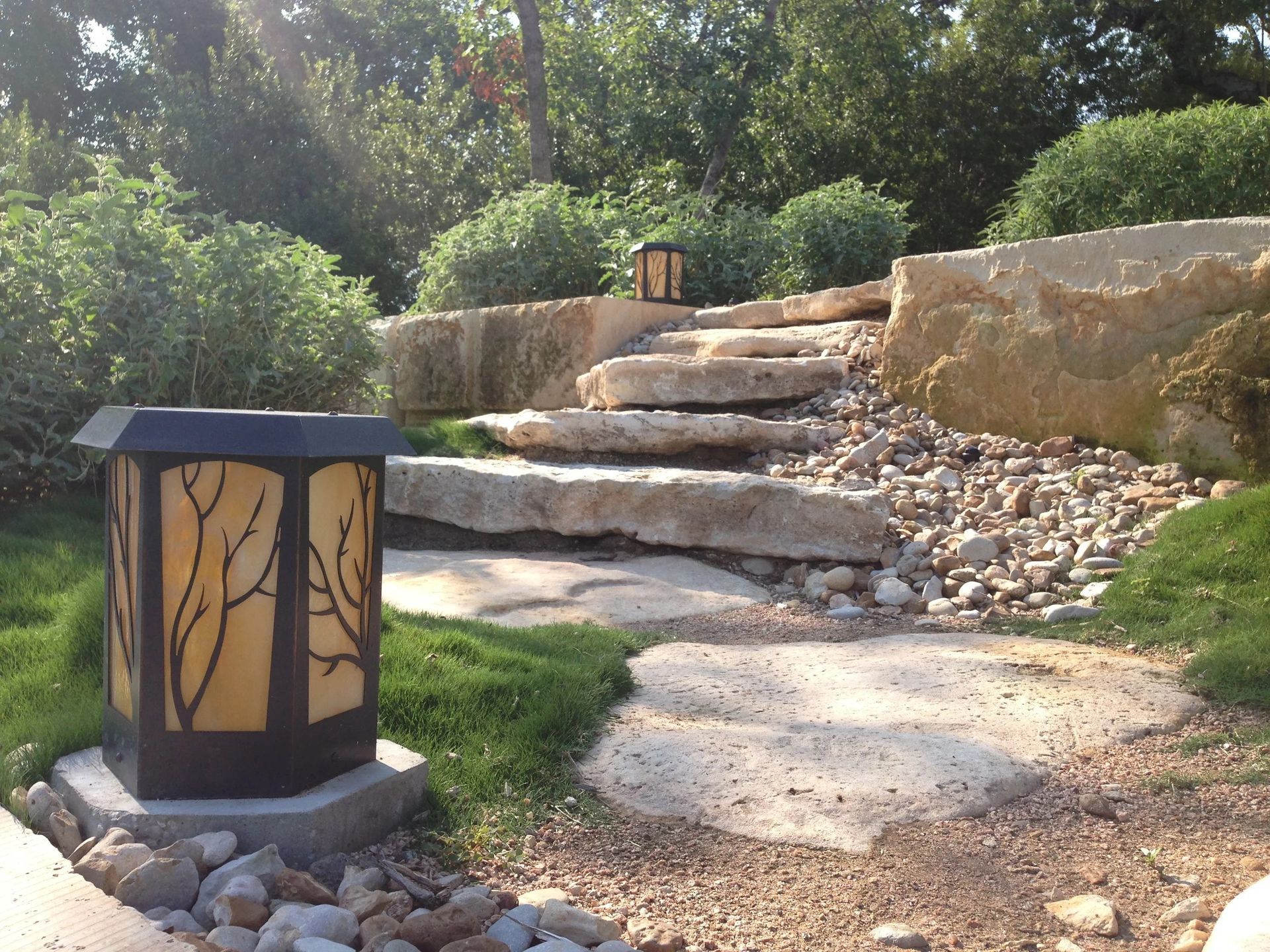 A garden path with large stone steps, surrounded by green shrubs, gravel, and an ornate, branch-patterned lantern.