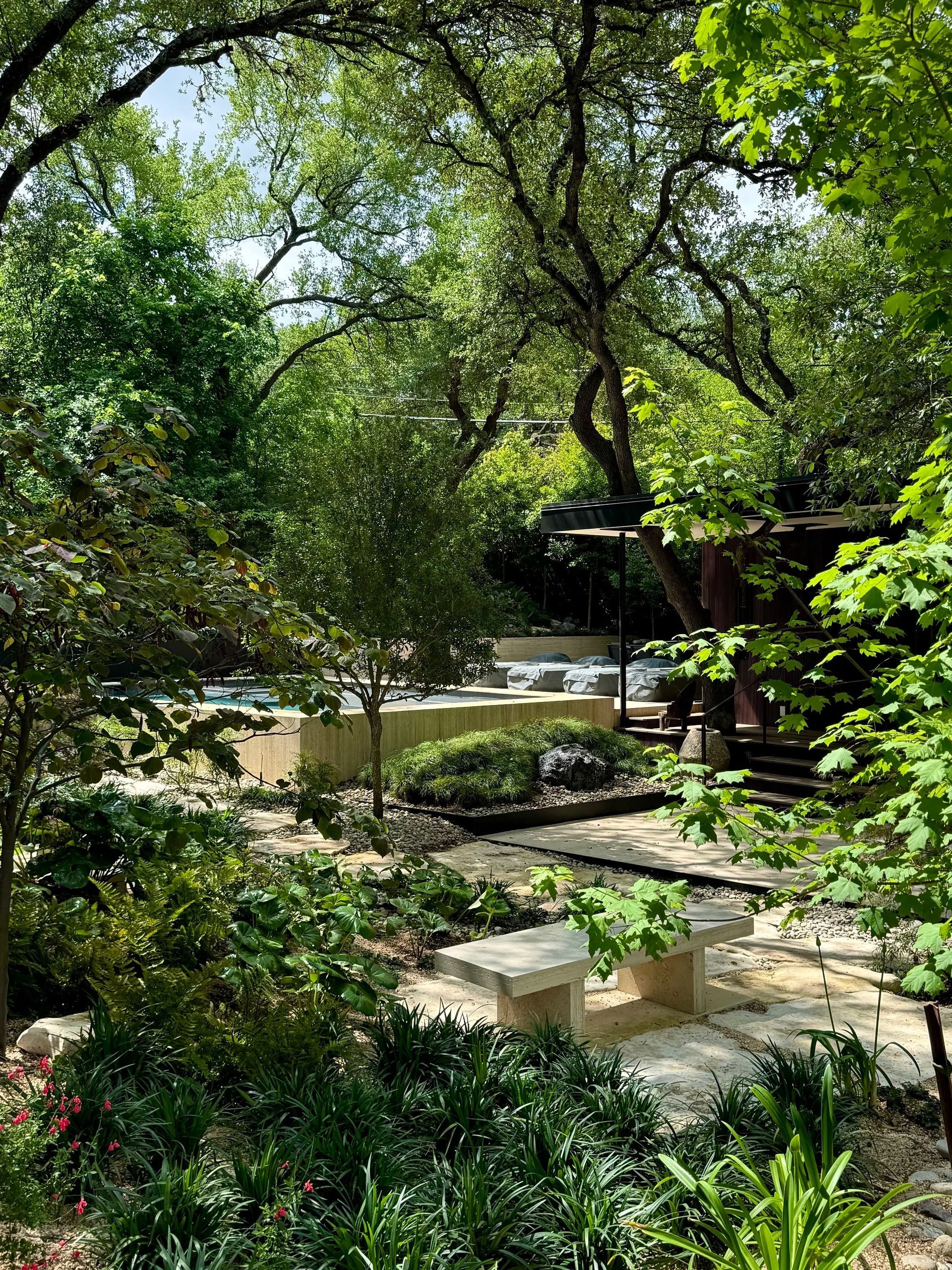 Stone bench in a lush garden, with a pool and trees in the background.