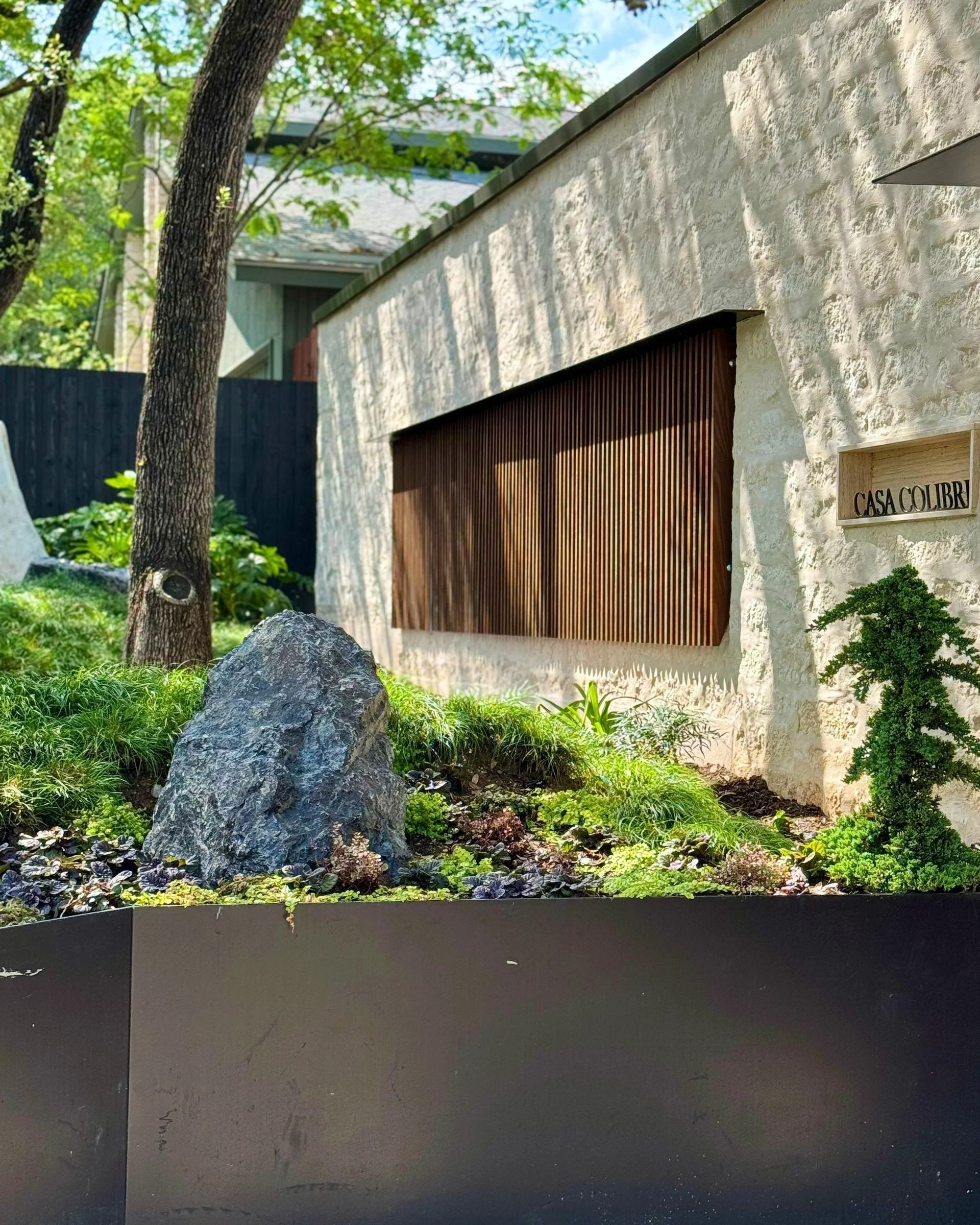 Exterior of building with textured white wall, wooden slats, and landscaped garden with rock.