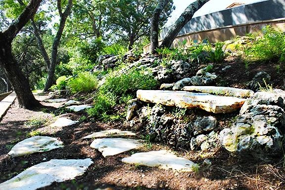 A garden pathway with flagstone stepping stones leading toward a stone bench on a wooded, terraced hillside.