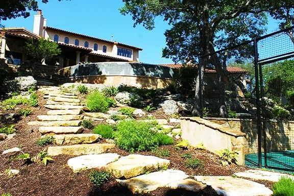 A landscape featuring a stone staircase leading up a hill to a Mediterranean-style home near a fenced outdoor court.