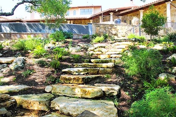 Large, rough-cut stone steps lead up a terraced garden toward a house with a stone patio and wall.