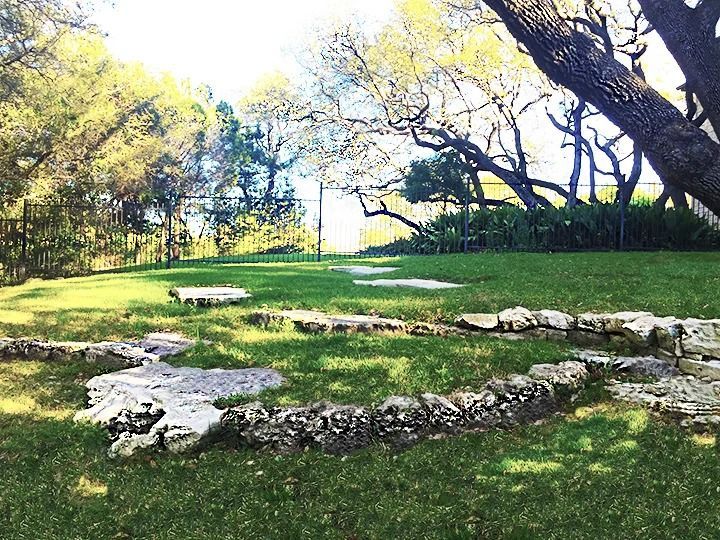 An outdoor scene featuring a stone path and rock border set in a grassy yard under large, sunlit trees.