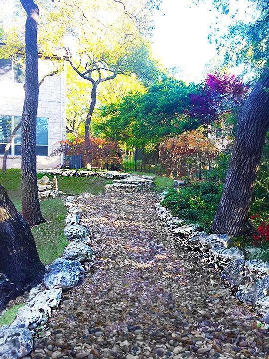A gravel path winds through a residential yard lined with decorative rocks, trees, and colorful bushes.