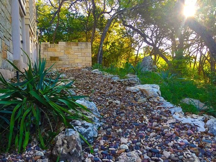 A gravel path winds through a garden with stone borders and a low stone wall, lit by sun filtering through trees.